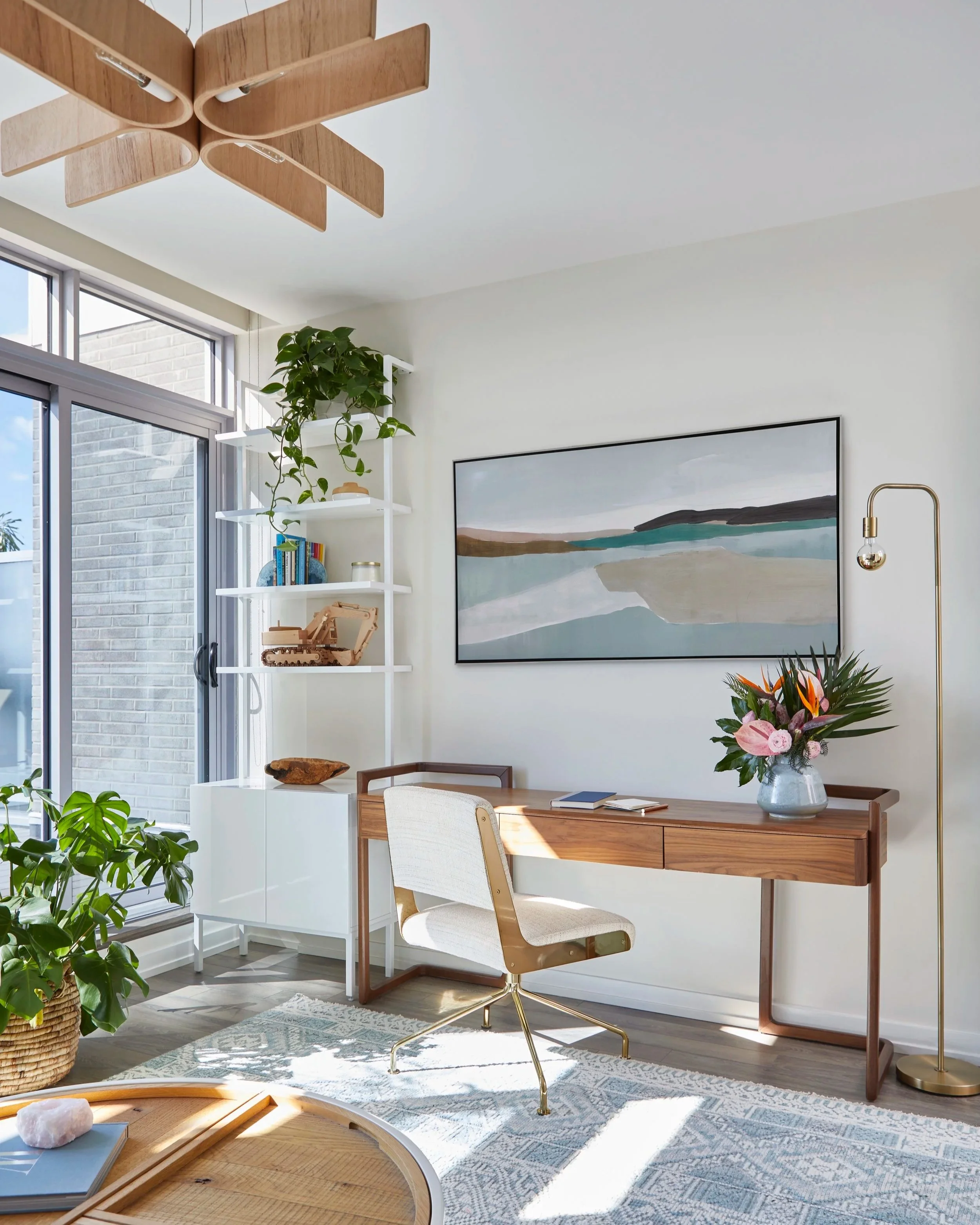 A bright home office with a wooden desk, beige chair, large abstract landscape painting, green plants, and a modern floor lamp.