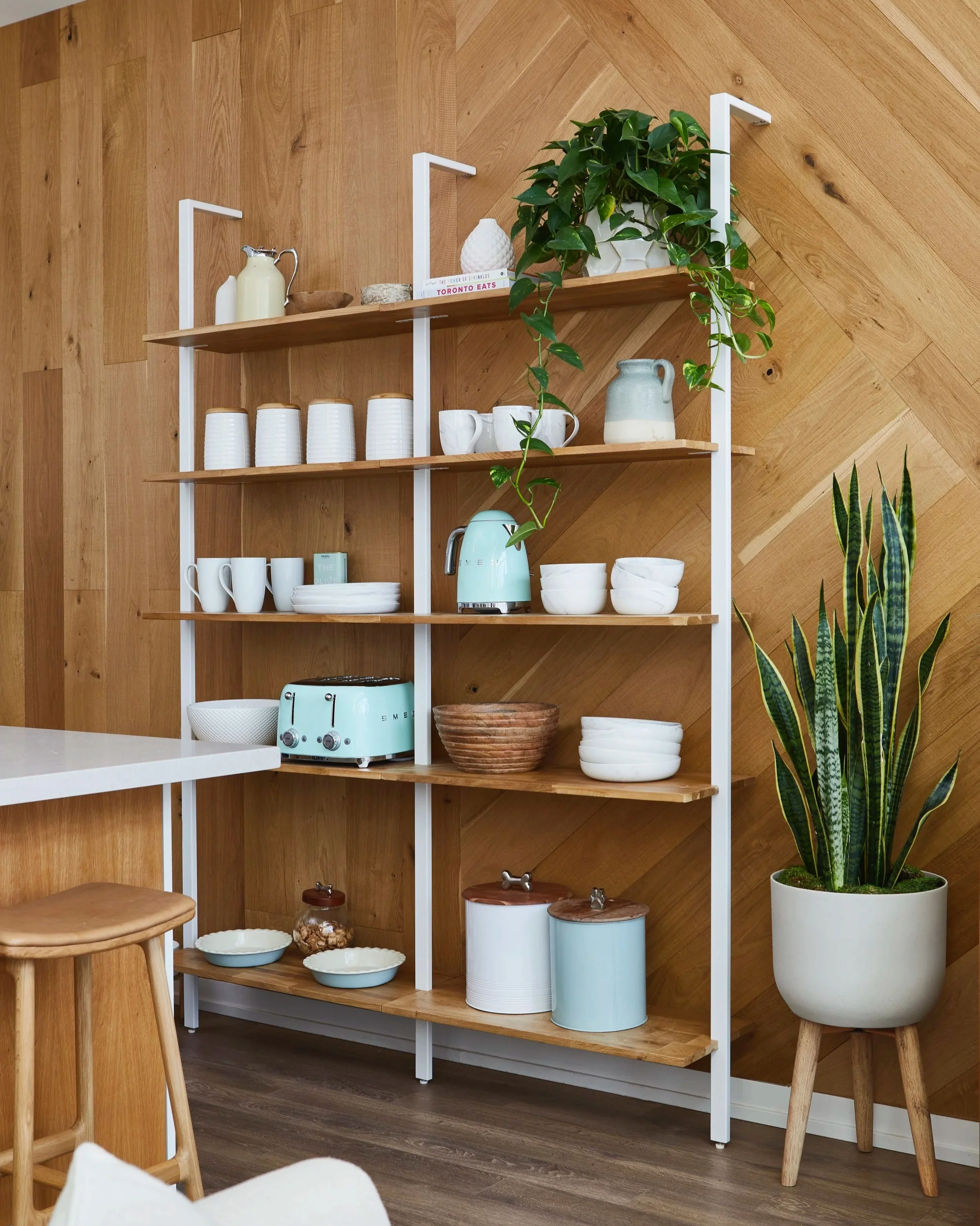 Wooden and white kitchen shelf with cups, bowls, a teal kettle, a teal toaster, and potted plants against a wood-paneled wall.