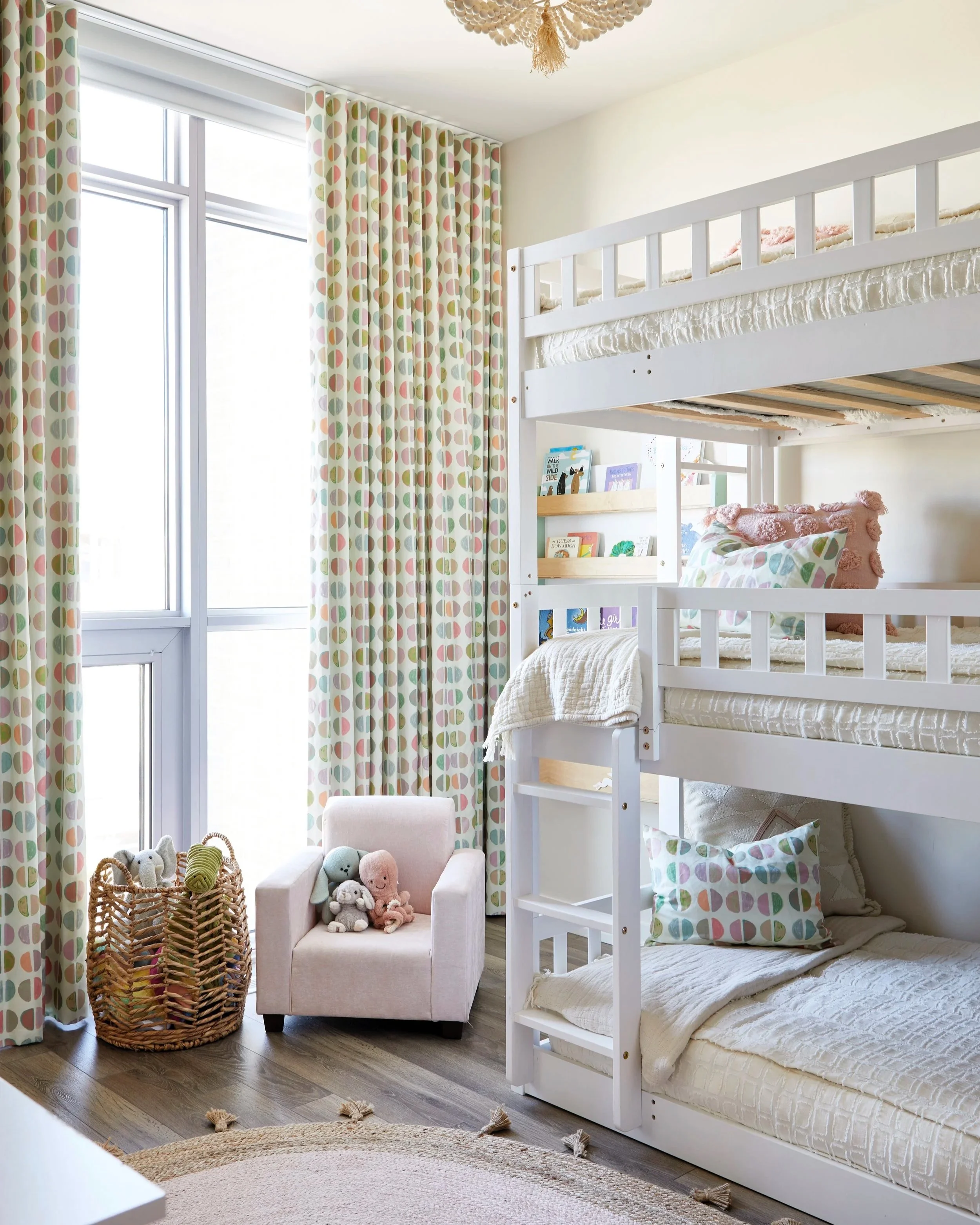 A bedroom with a white bunk bed, colorful patterned curtains, a small pink armchair with plush toys, and a woven basket filled with plush toys near a large window.