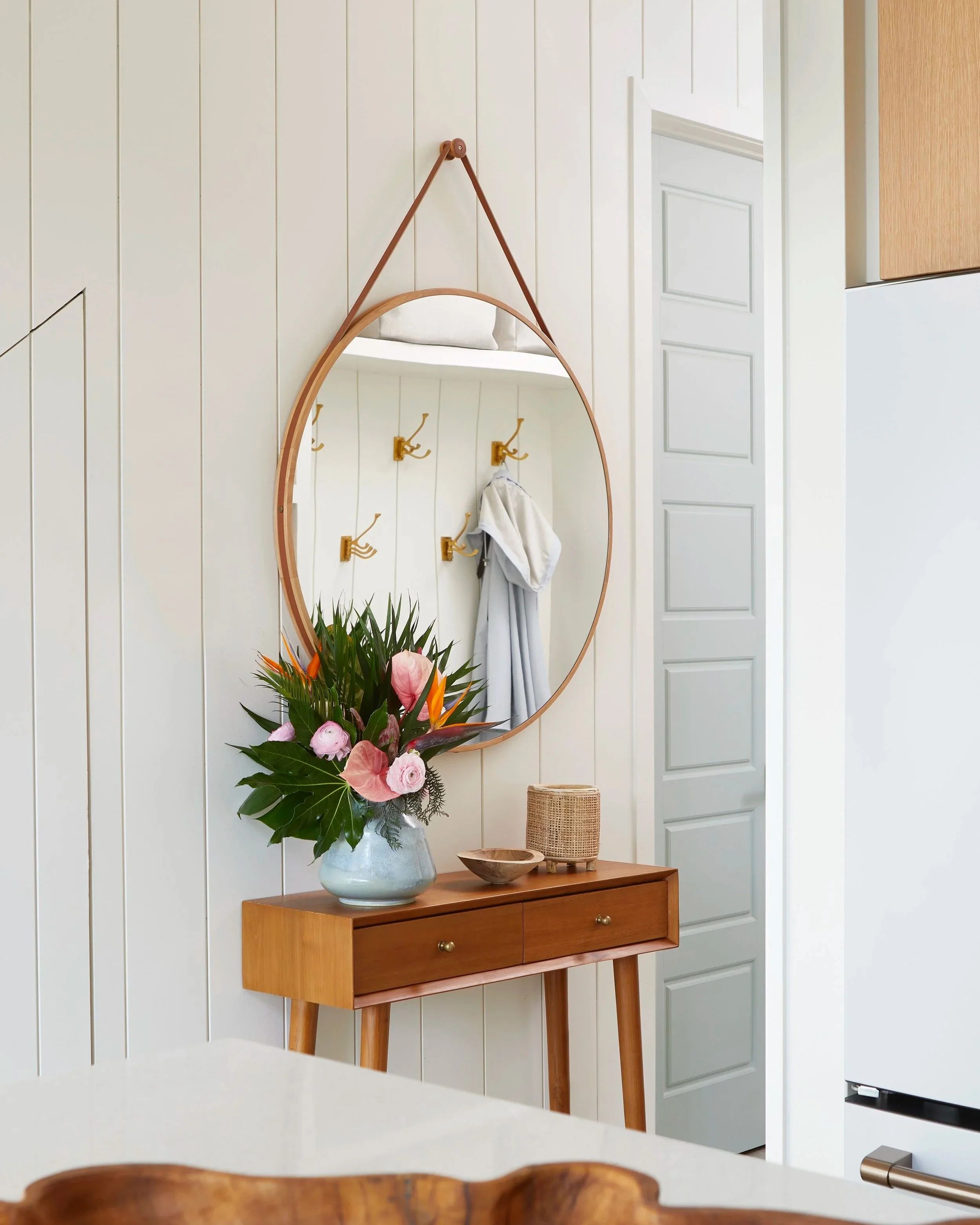 A wooden side table with a floral arrangement, a woven basket, and a wooden bowl in front of a large oval mirror on a white wall.