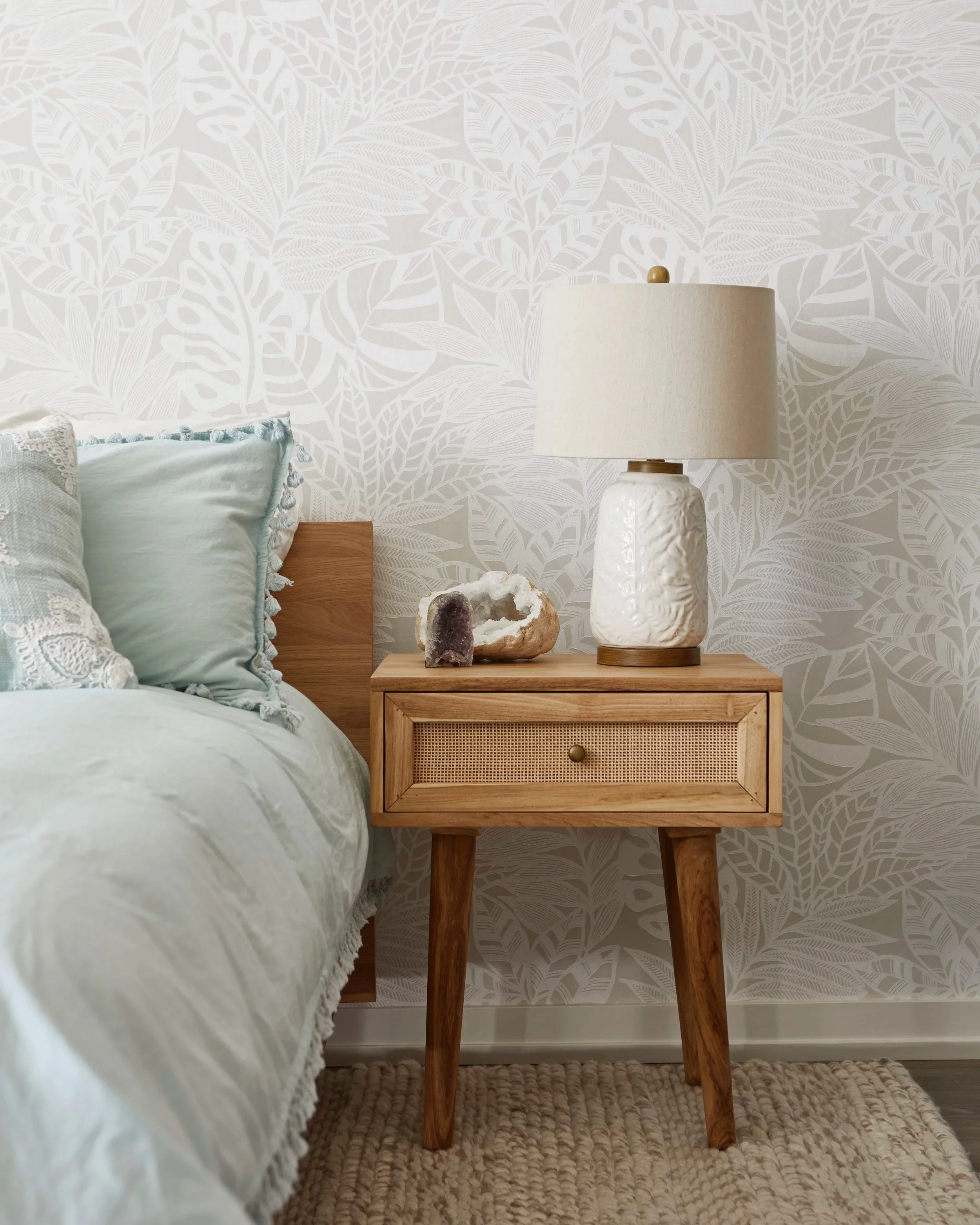 A bedroom scene with a wooden nightstand next to a bed, featuring a white textured lamp, a geode, and a decorative rock. The bed has light-colored bedding and pillows, and the background has a leafy wallpaper pattern.