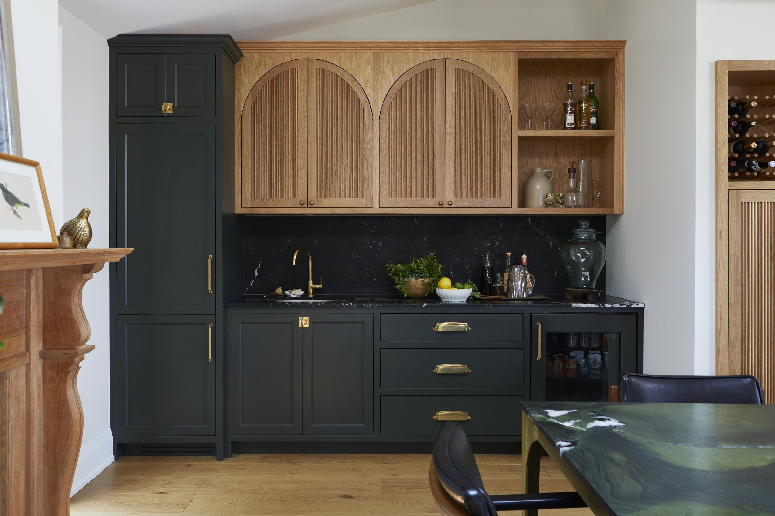 Modern kitchen with dark green lower cabinets, light wooden upper cabinets with arched design, black marble backsplash, and gold hardware. Kitchen counter has a gold faucet, potted herbs, lemon, and liquor bottles. Wooden floors and a dining table wi