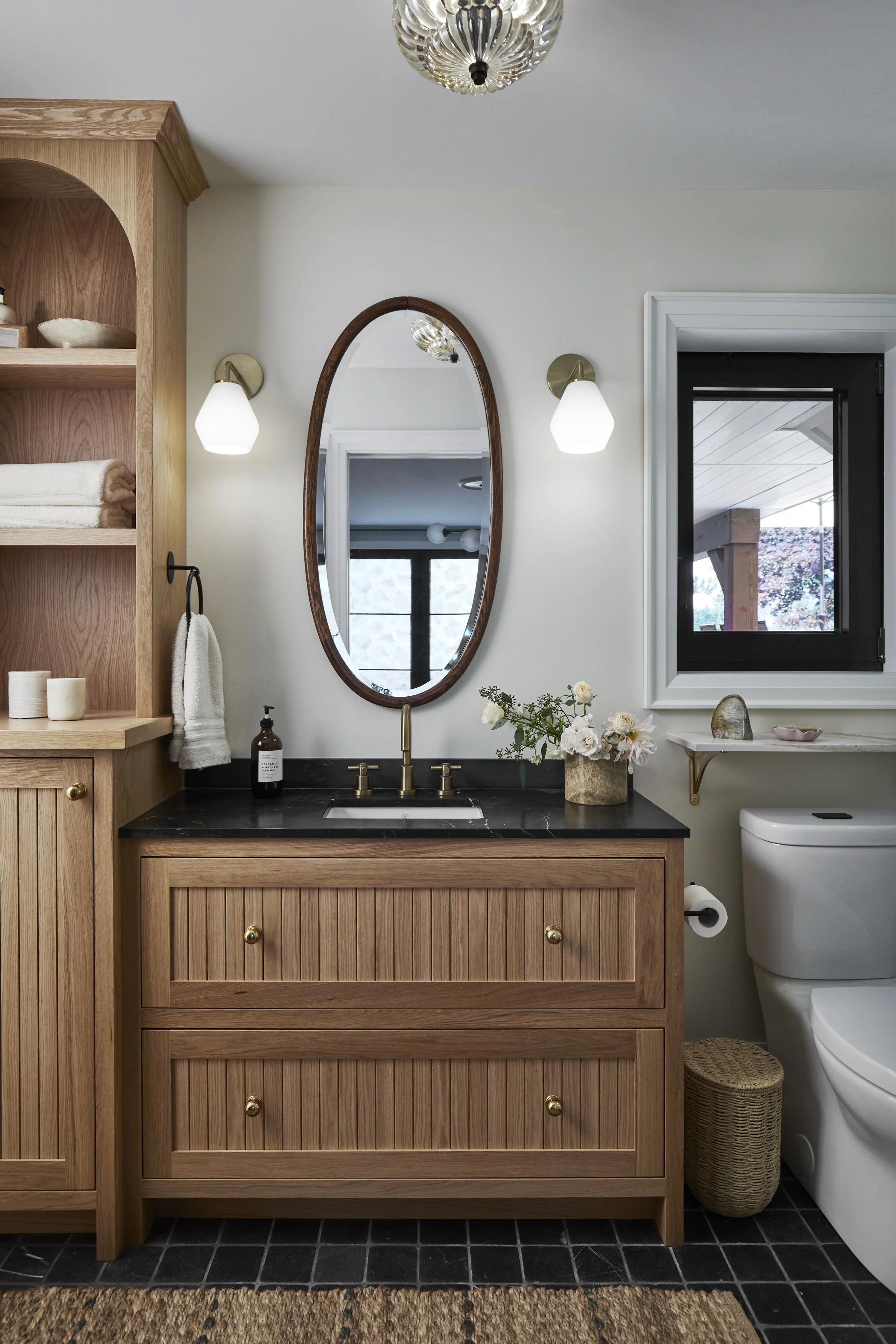 A bathroom featuring a wooden vanity with a black countertop, an oval mirror, two wall sconces, and a window. There are decorative items such as a vase with flowers and small dishes, with a toilet nearby.
