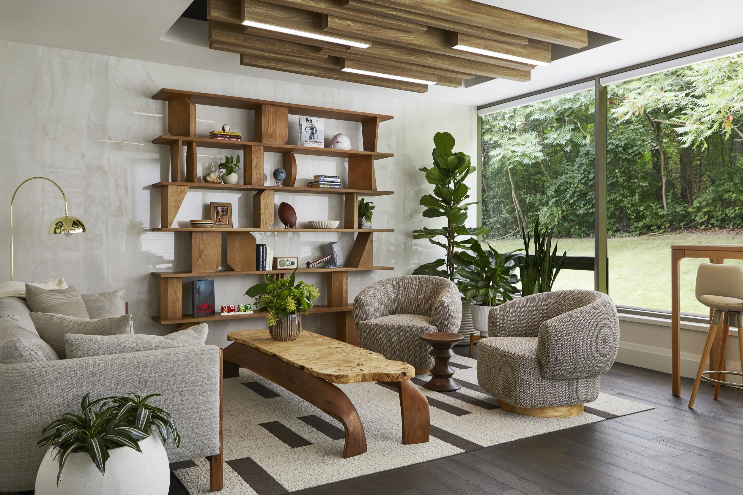 A modern living room with beige sofa, two gray armchairs, a wooden coffee table with a natural edge, a white patterned rug, and a large window revealing a lush green outdoor scene. The room features a wooden ceiling design, a wall-mounted bookshelf w