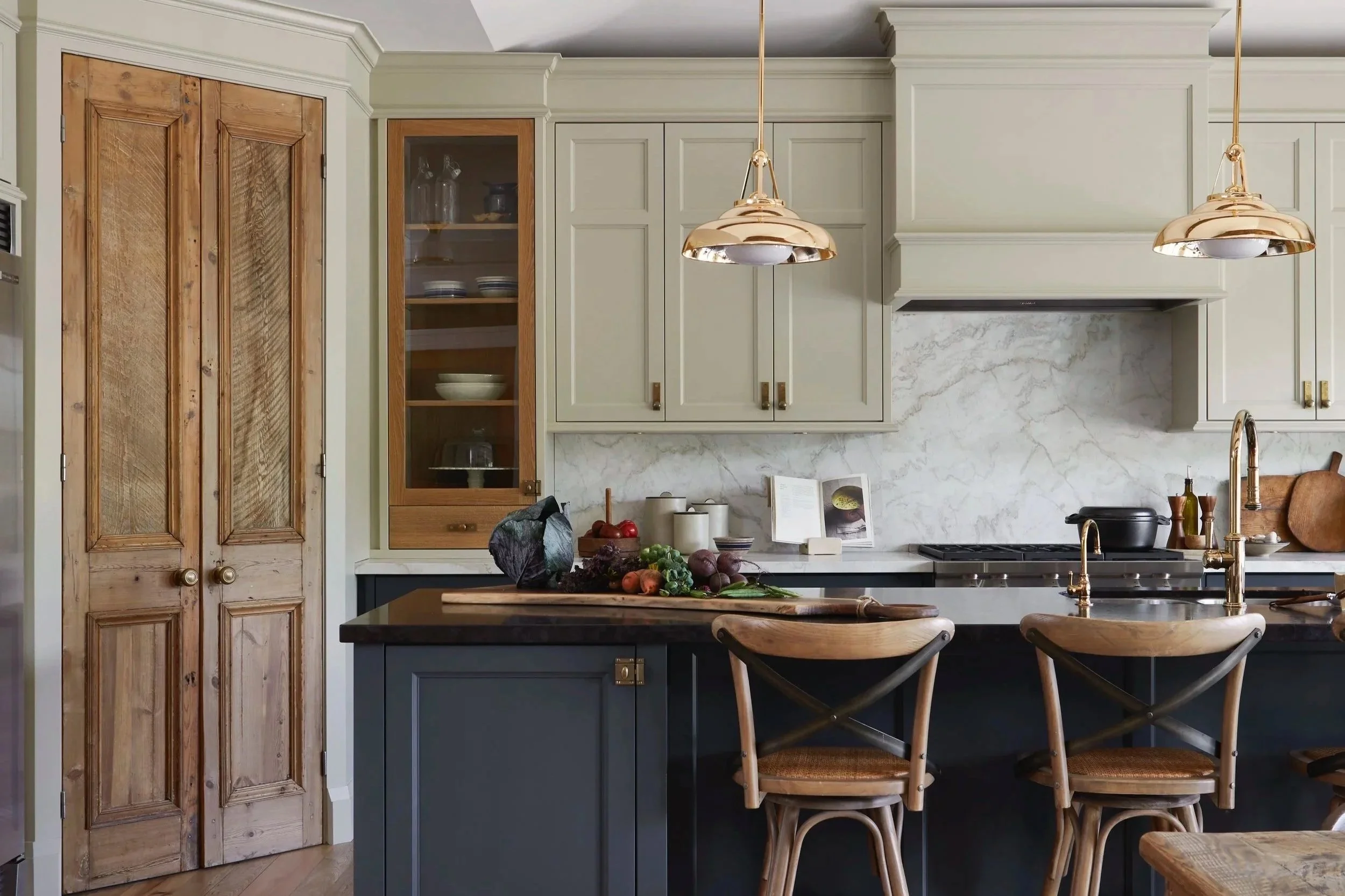 Kitchen with beige cabinets, a black island countertop, and wooden chairs. There are copper pendant lights hanging above the island, and a marble backsplash and countertop. Various vegetables and a book are on the counter.
