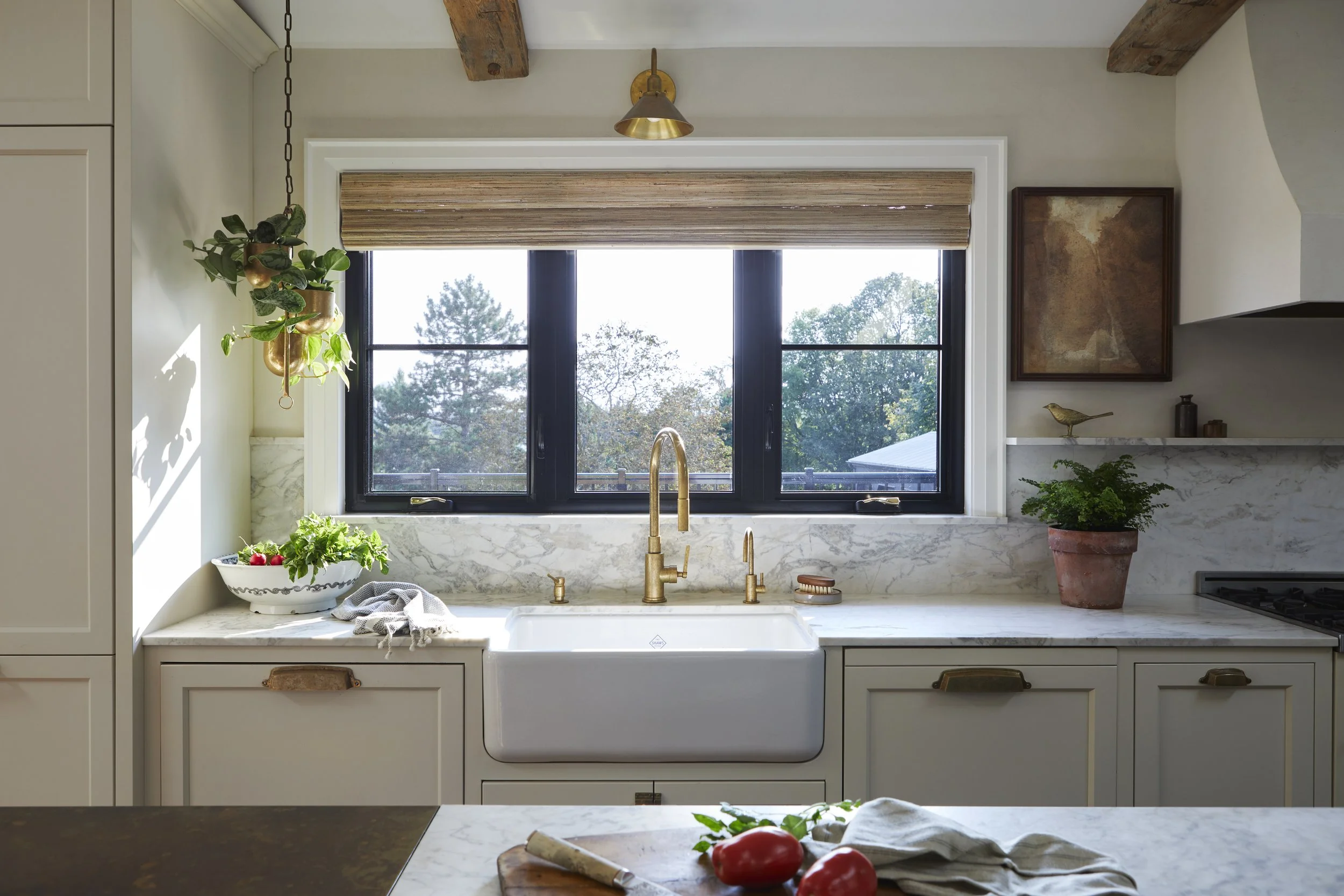 Modern kitchen with large window above farmhouse sink, brass faucet, potted plants, fresh vegetables, and natural decor.