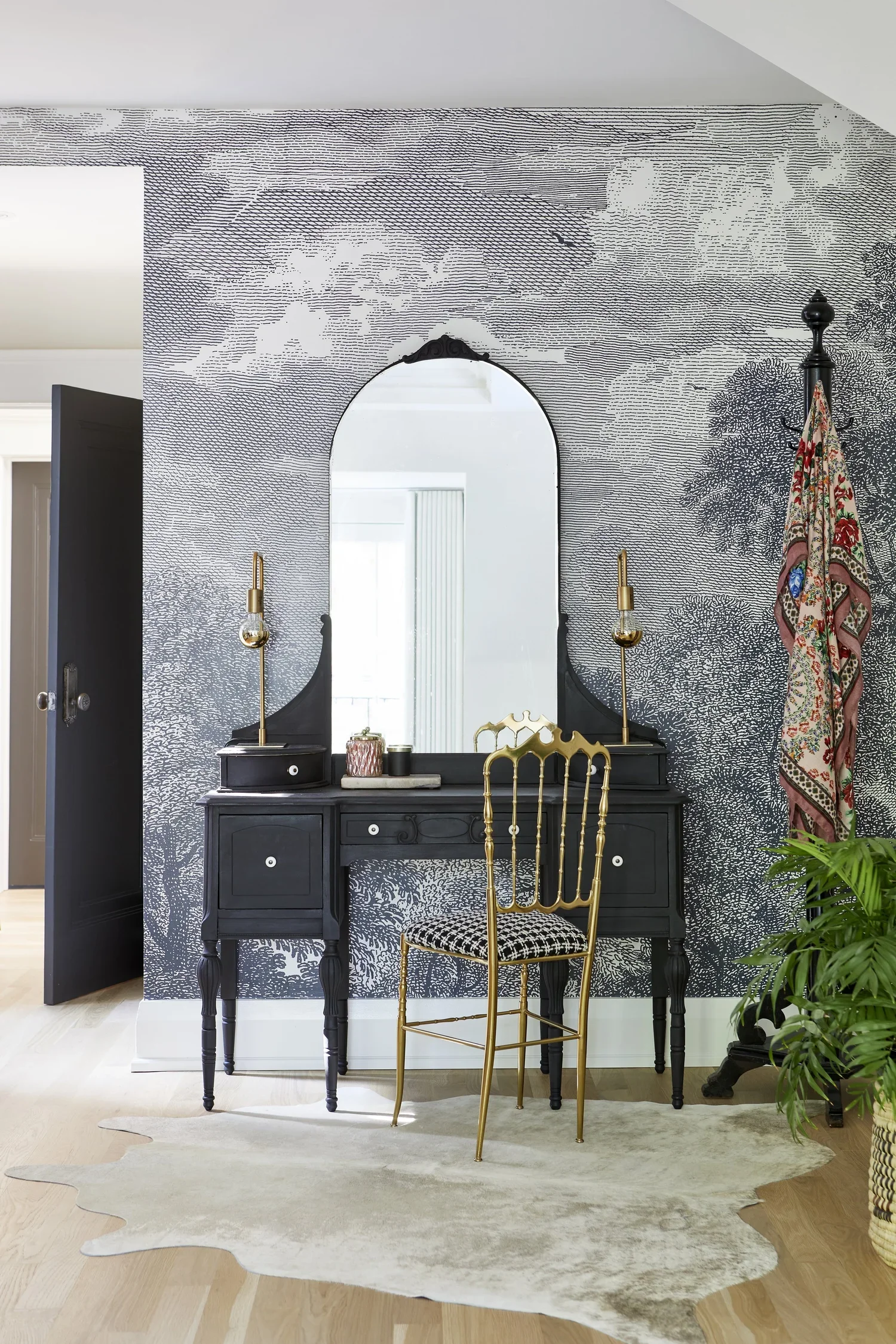 A black vanity desk with a mirror, gold accents, and two table lamps against a black and white nature-themed wall mural. A gold chair with a black and white patterned cushion is in front of the desk. There is a cowhide rug on the wooden floor, and a 