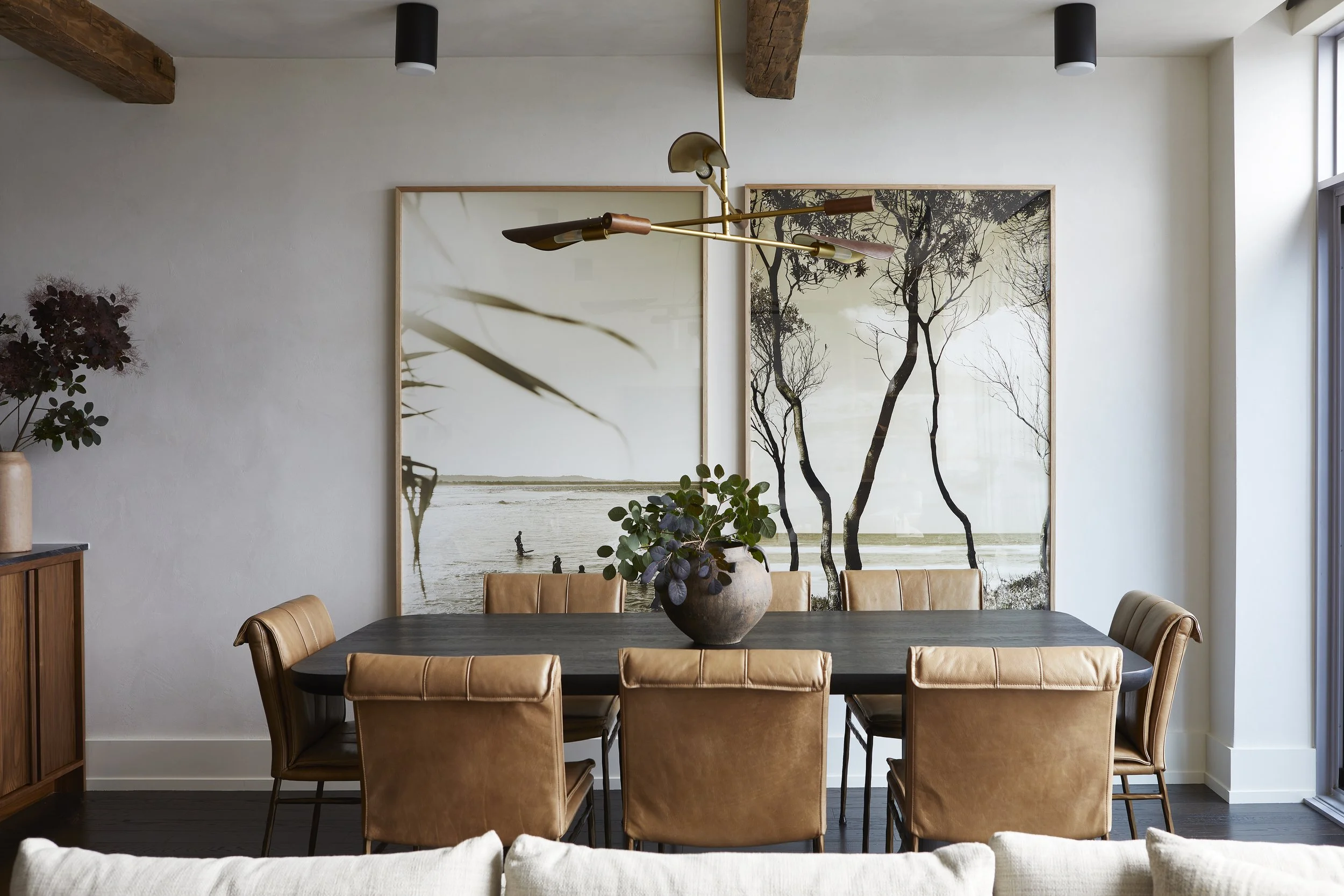 Modern dining room with a black table, tan leather chairs, and large black-and-white framed artwork of trees and a beach scene on the wall. There's a decorative plant in a round vase on the table, a wooden sideboard on the left, and a gold and black modern chandelier hanging above.