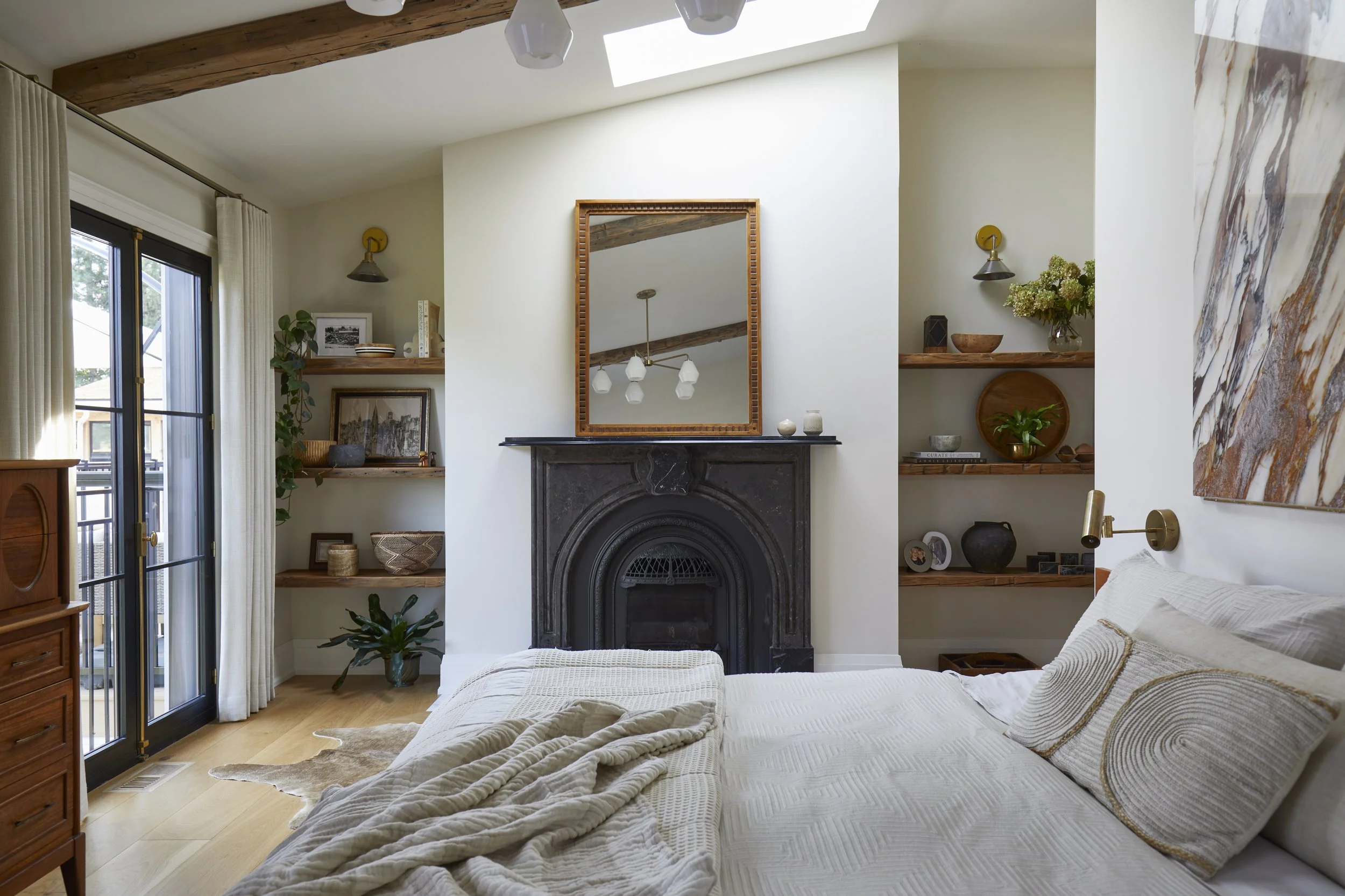 Cozy bedroom with large bed, cream bedding, wooden shelves with decorative items, a black fireplace with a mirror above, a large abstract painting, and a sliding glass door leading to a balcony.