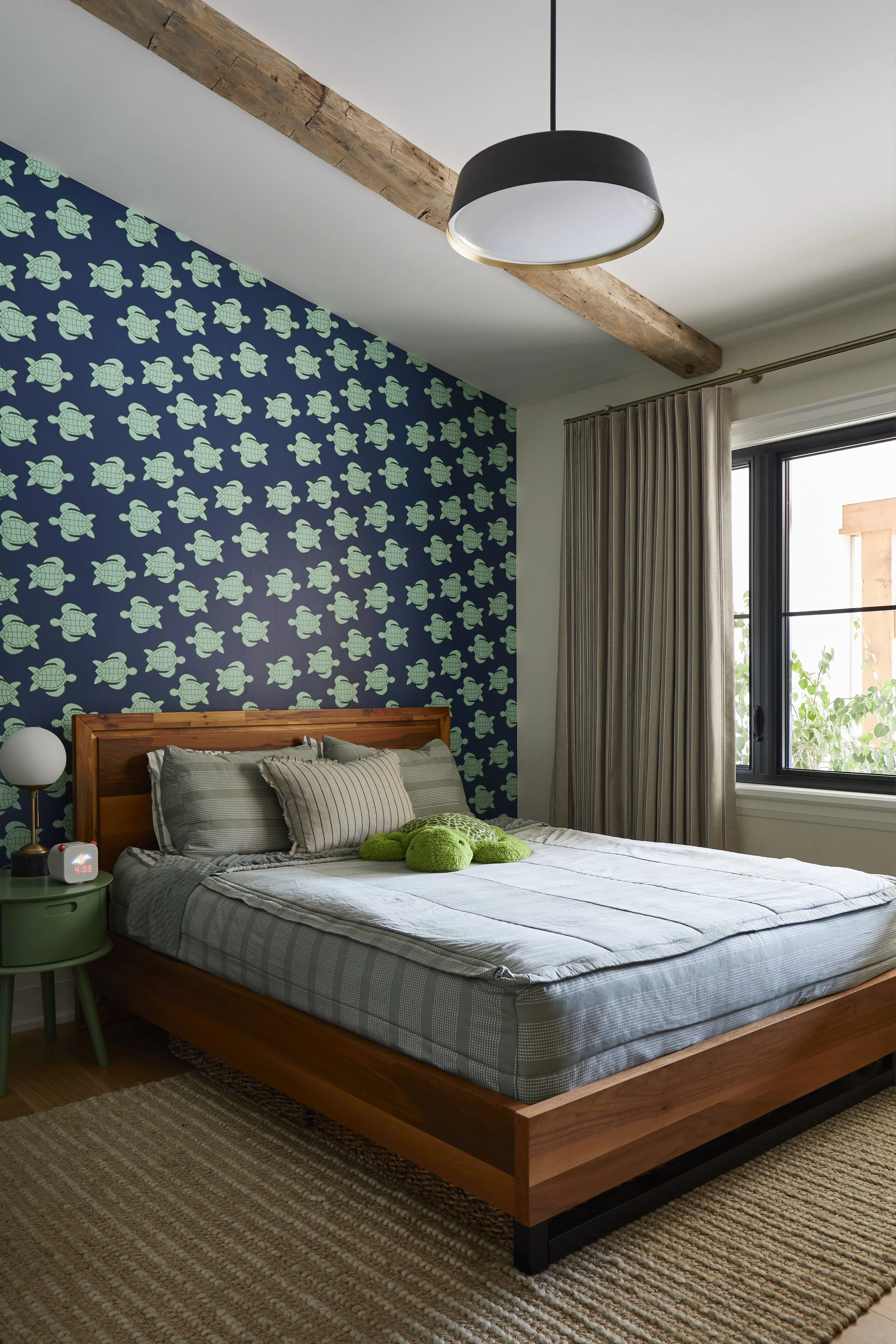 A bedroom with a wooden bed frame, dressed with striped gray and white bedding, and a green plush turtle toy on top. The wall behind the bed has blue wallpaper with green turtle patterns. To the left, a green nightstand with a white lamp and alarm cl
