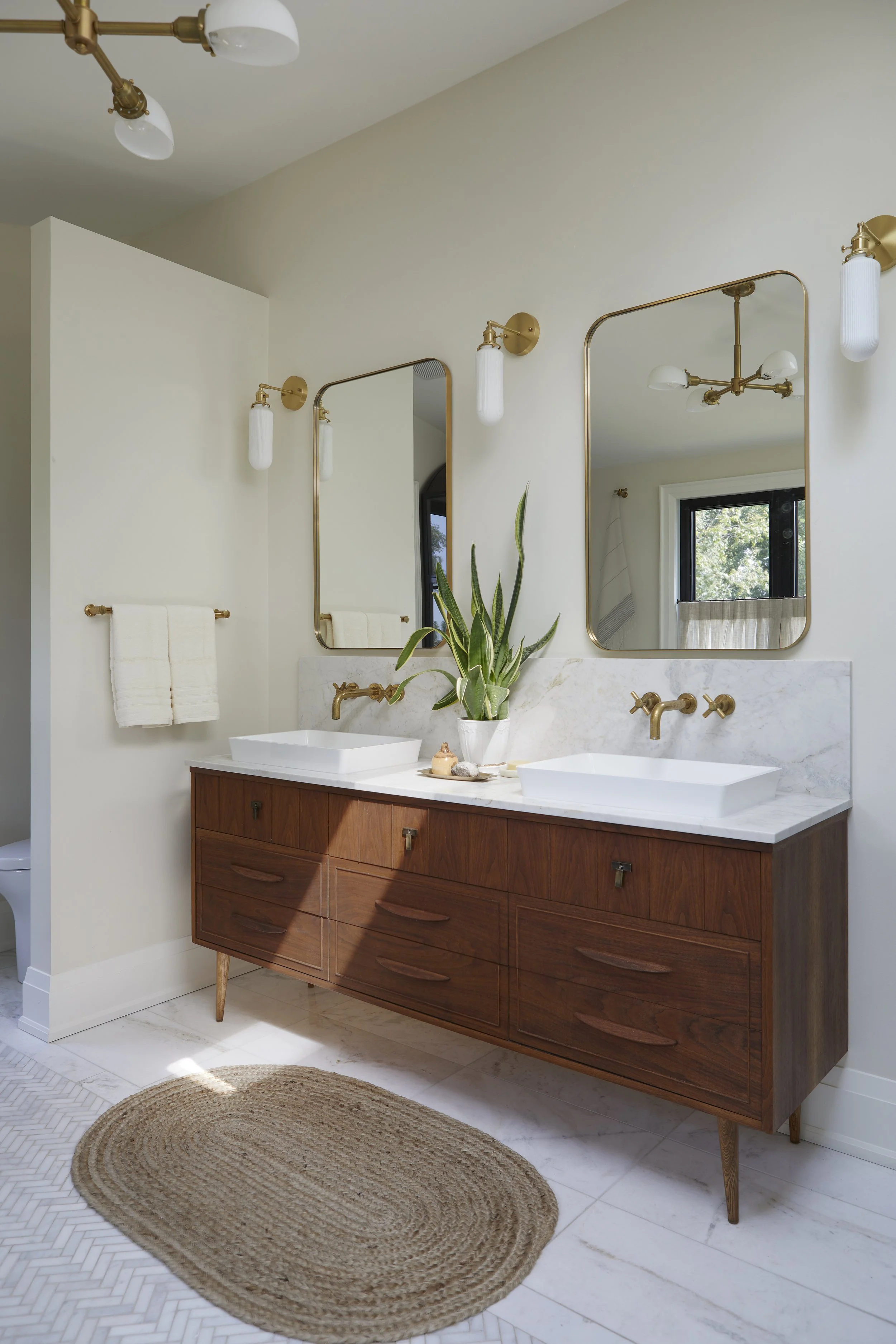 A modern bathroom featuring a wooden vanity with two vessel sinks, three mirrors, brass fixtures, wall-mounted light fixtures, a potted plant, a towel rack with white towels, a window with a curtain, and a beige rug on white tile flooring.
