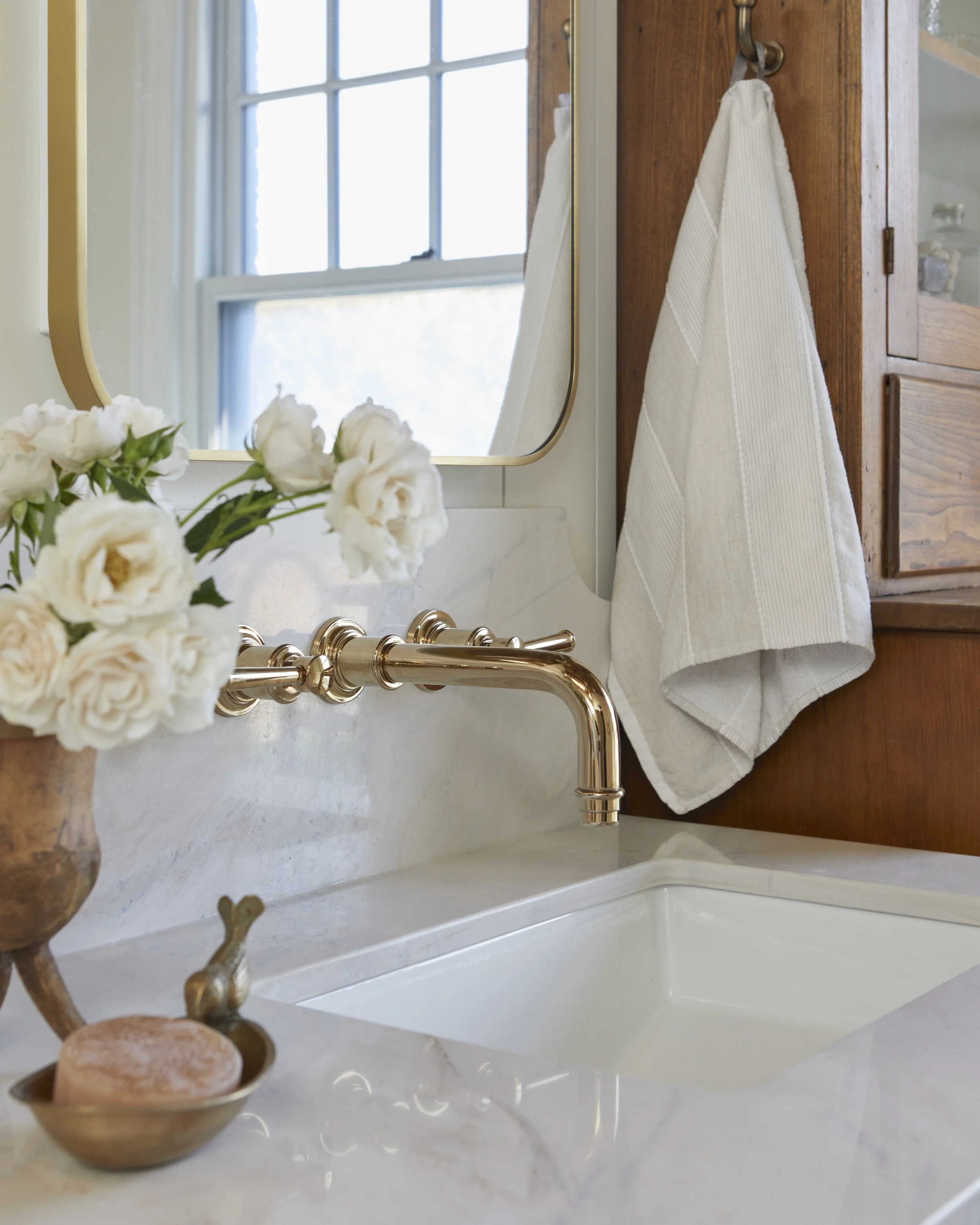 A bathroom sink with a white marble countertop, a vase of white flowers, a brass faucet, a mirror, and a towel hanging on a wooden cabinet door.