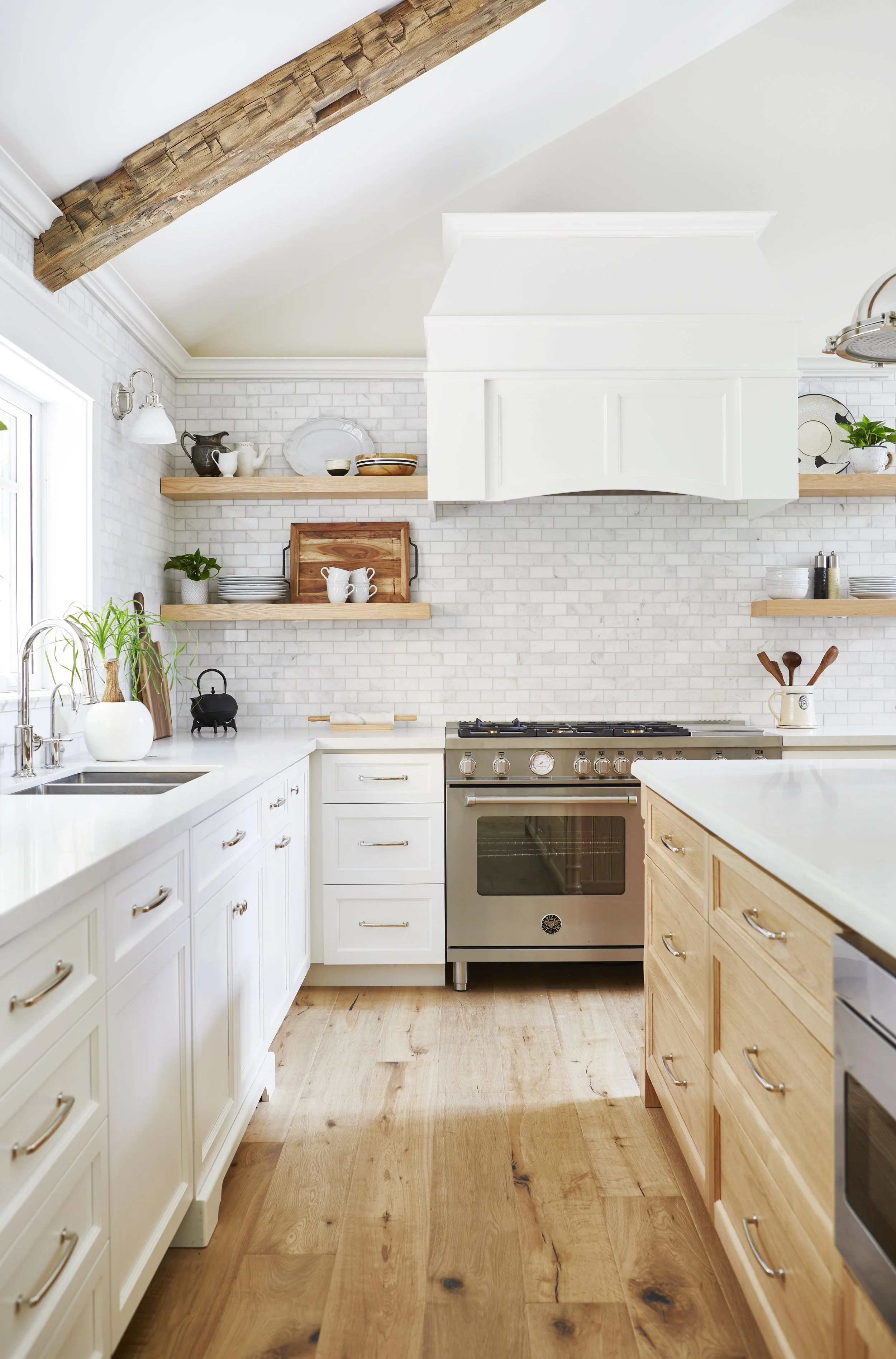A bright kitchen with white cabinets, a stainless steel stove, open wooden shelves with dishes, and a wooden beam on the ceiling.