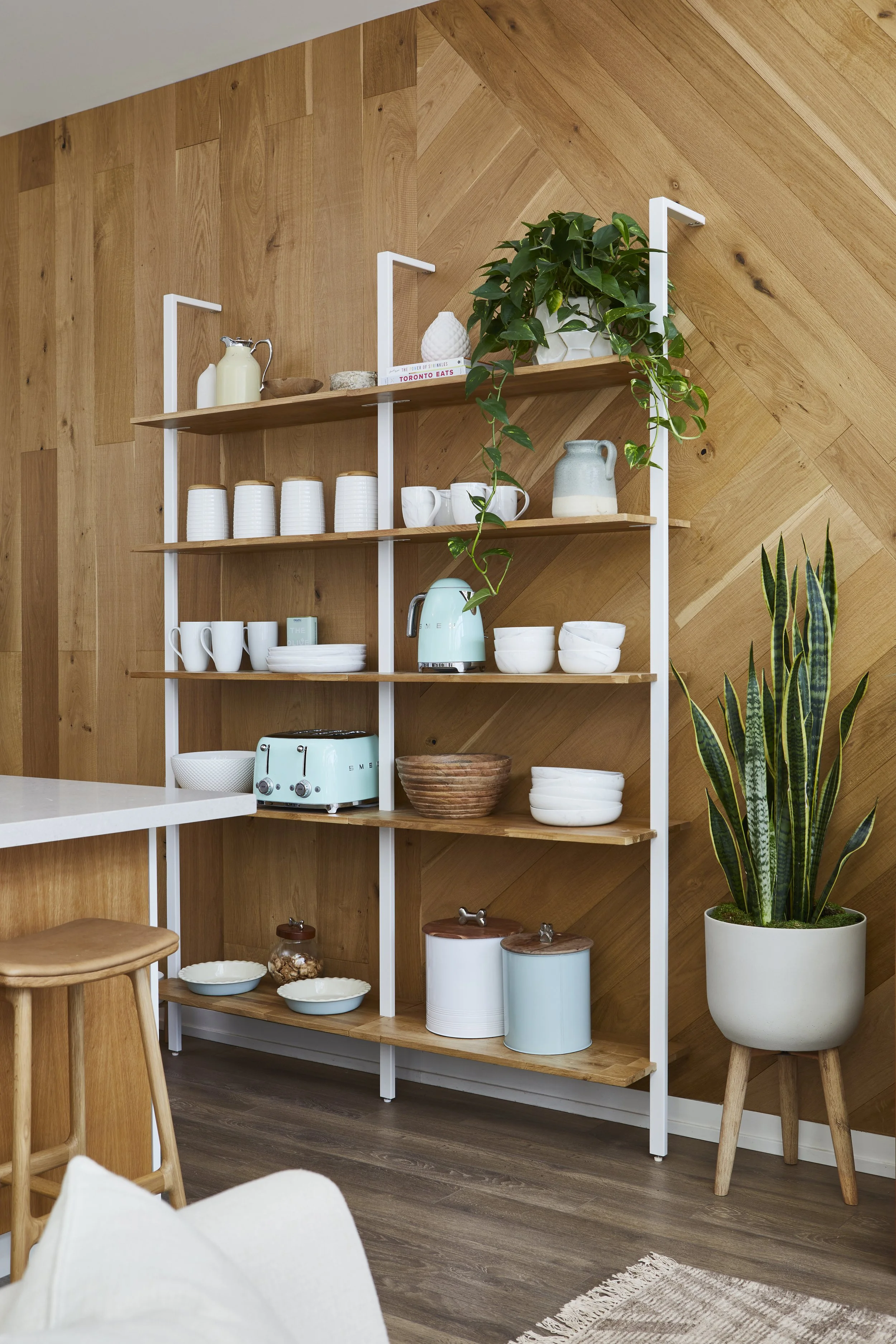 Open wooden shelving unit with kitchenware and decor, next to a wooden wall and potted snake plant.
