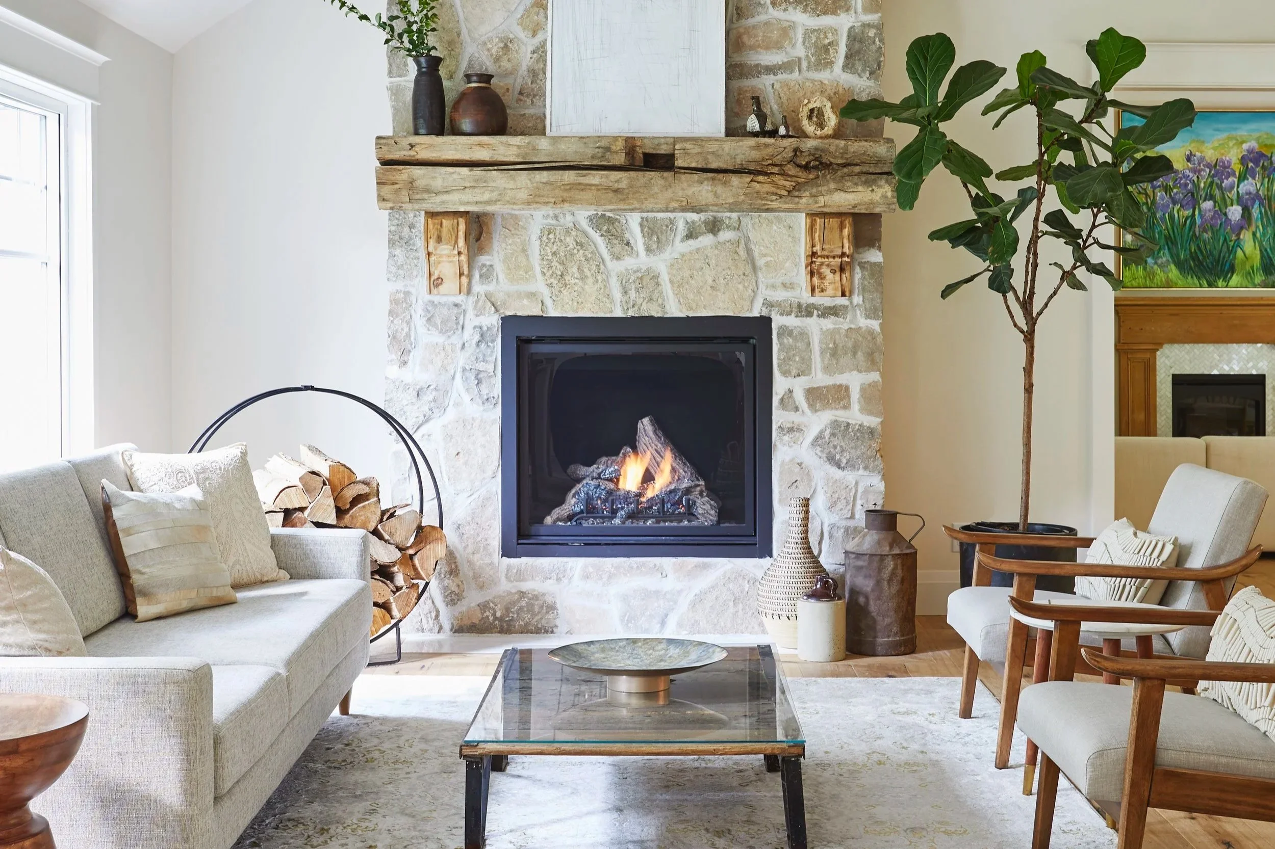 Living room with a stone fireplace, wooden mantel, and a fire burning. To the left, a beige sofa with pillows, and on the right, a wooden armchair with cream cushions. A glass-topped coffee table is in the center. There is a large potted tree, decorative vases, and artwork on the walls.