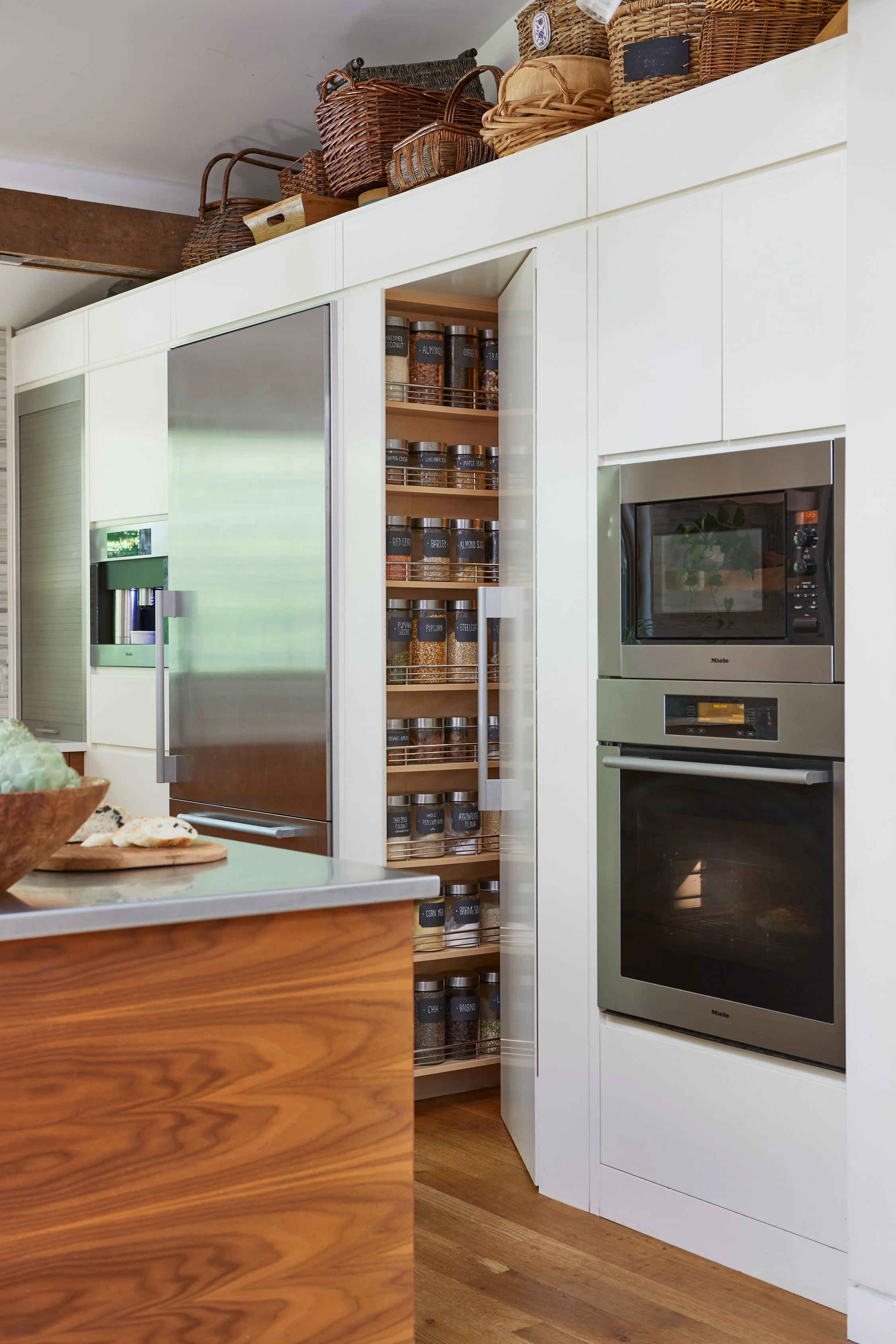 Kitchen with white cabinets, wooden shelf with glass jars of spices, stainless steel refrigerator, built-in microwave and oven, and baskets on top of cabinets.