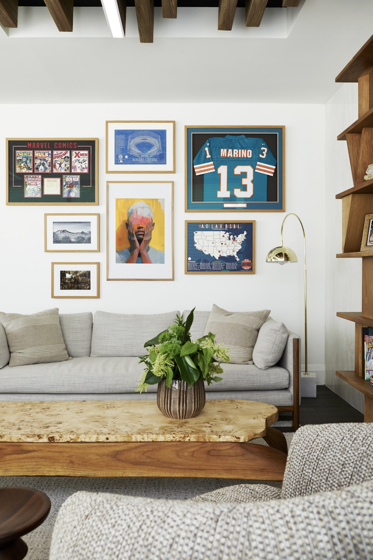 Living room with a beige sofa, a wooden coffee table with a green plant, and a wall decorated with framed sports memorabilia and artworks including a framed Miami Dolphins jersey, a map, and Marvel comic covers.