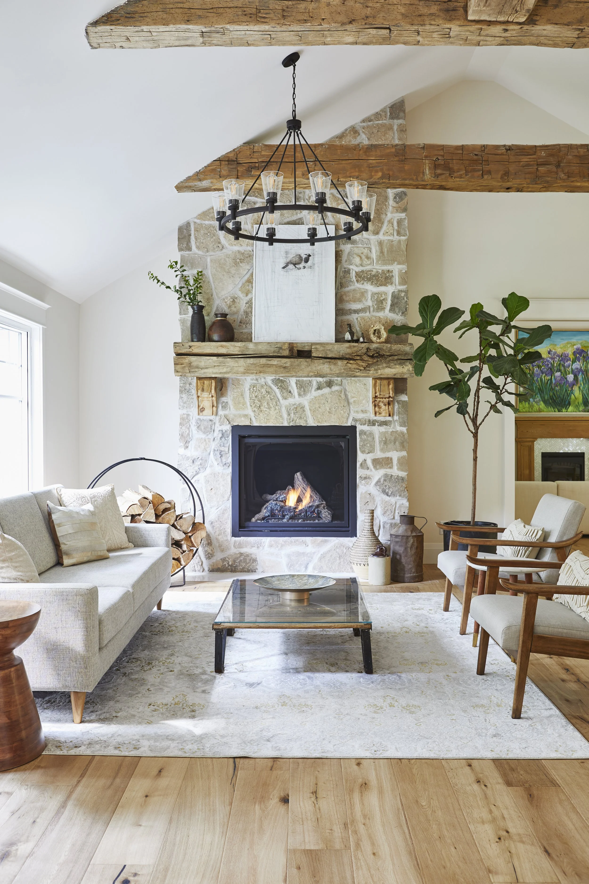 Living room with a stone fireplace, wood beams, light-colored sofa, armchairs, a glass coffee table, decorative vases, potted plant, chandelier, and a window.