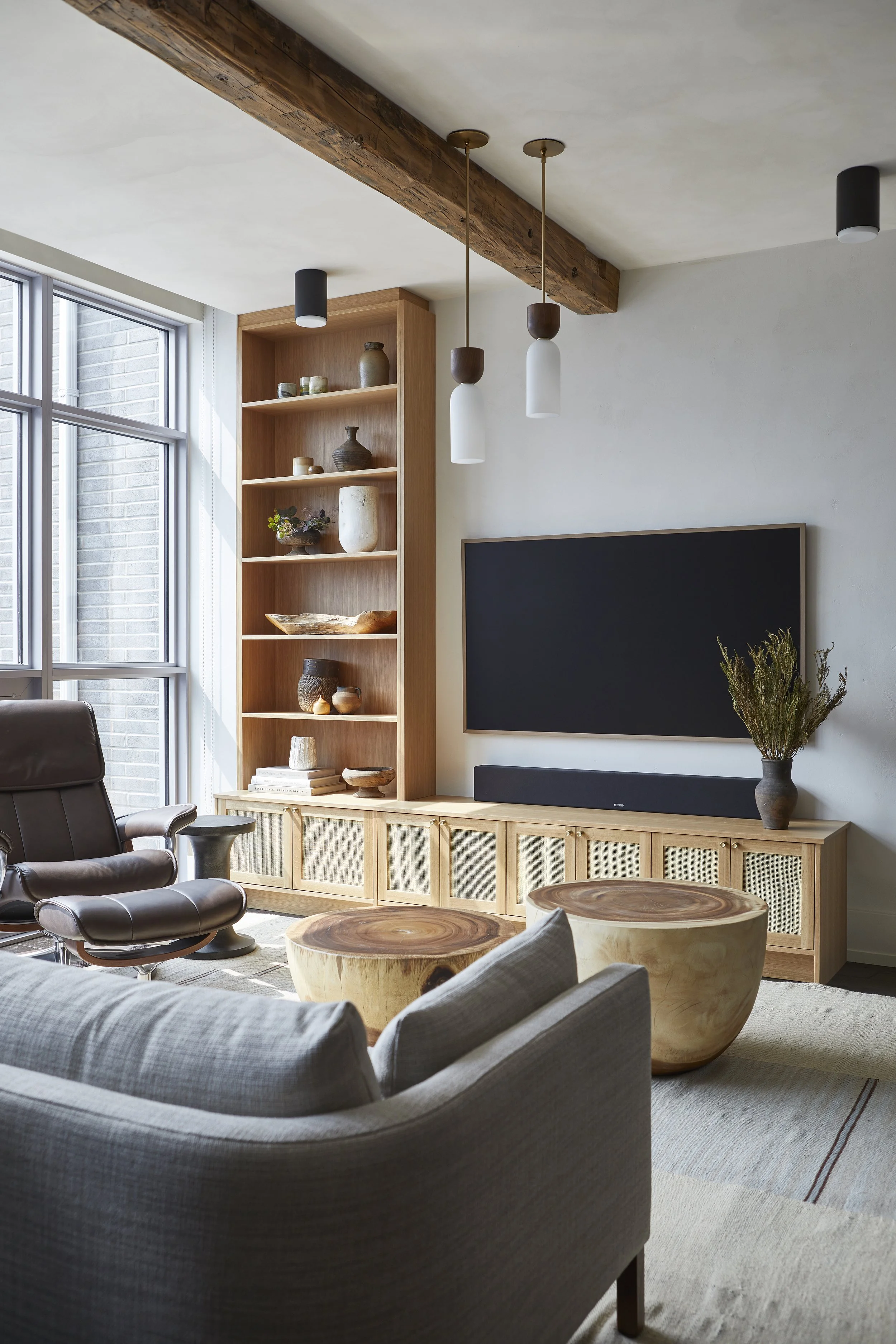 Living room with wooden shelving, a large flat-screen TV, a sofa, armchair, and two round wooden coffee tables, natural light from a large window, and modern decor.