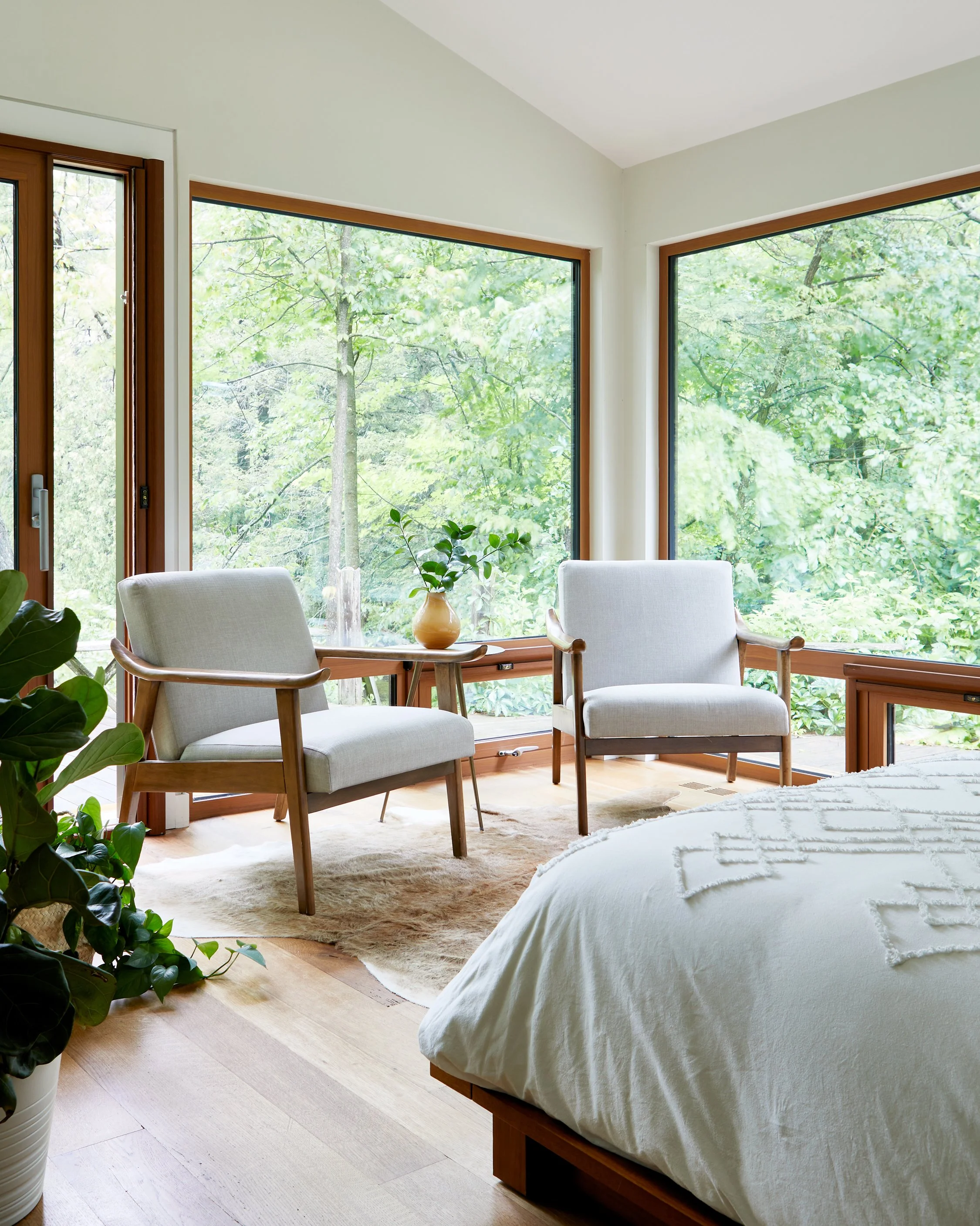 A bedroom with large windows overlooking a green wooded area, two beige armchairs, a small table with a plant, a bed with a white blanket, and a potted plant in the corner.