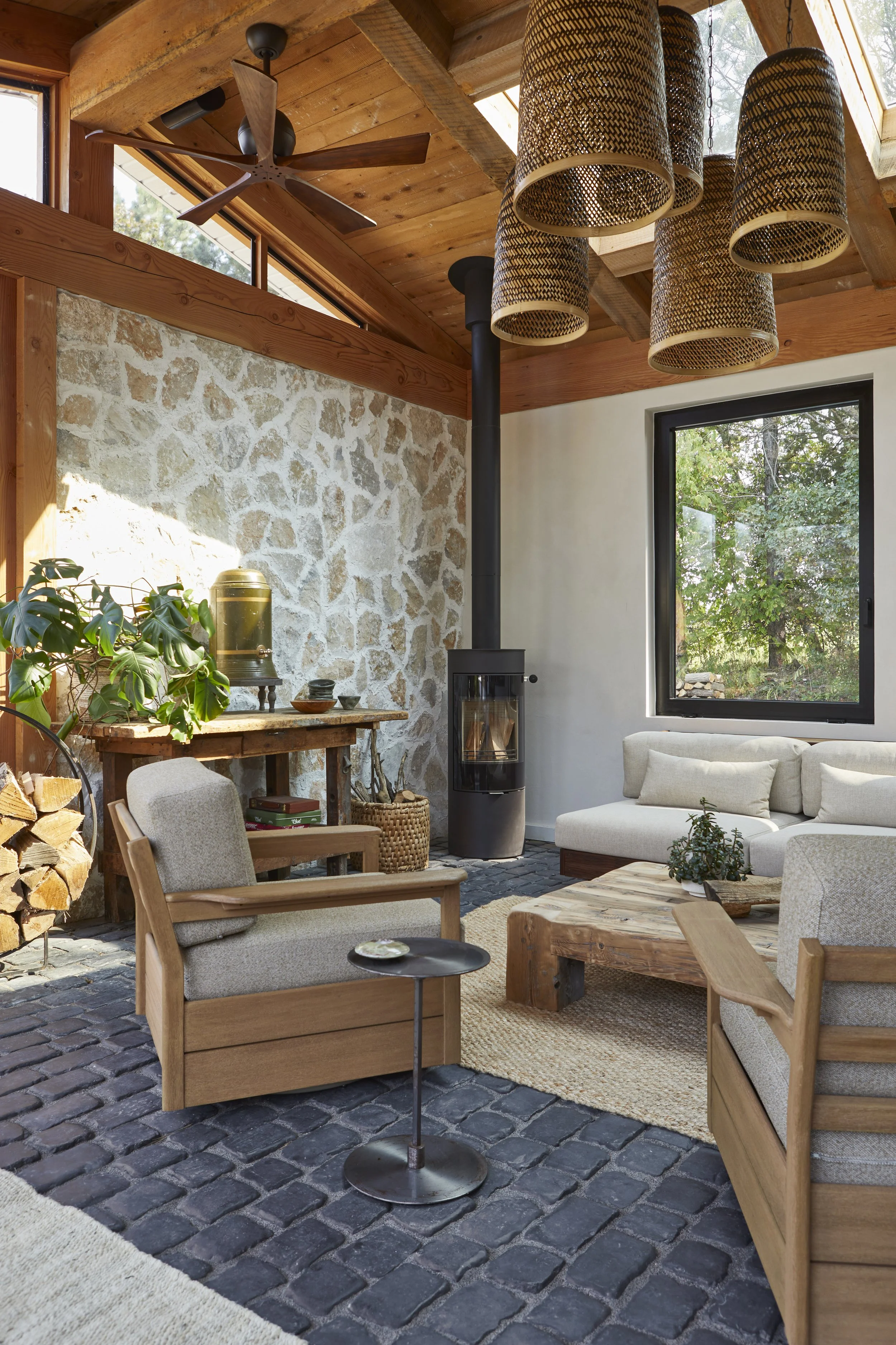 Cozy living room with natural wood ceiling, stone wall, and large black window, featuring light-colored sofa, wooden armchairs, a rustic coffee table, wood logs, a wood stove, and decorative hanging lights.