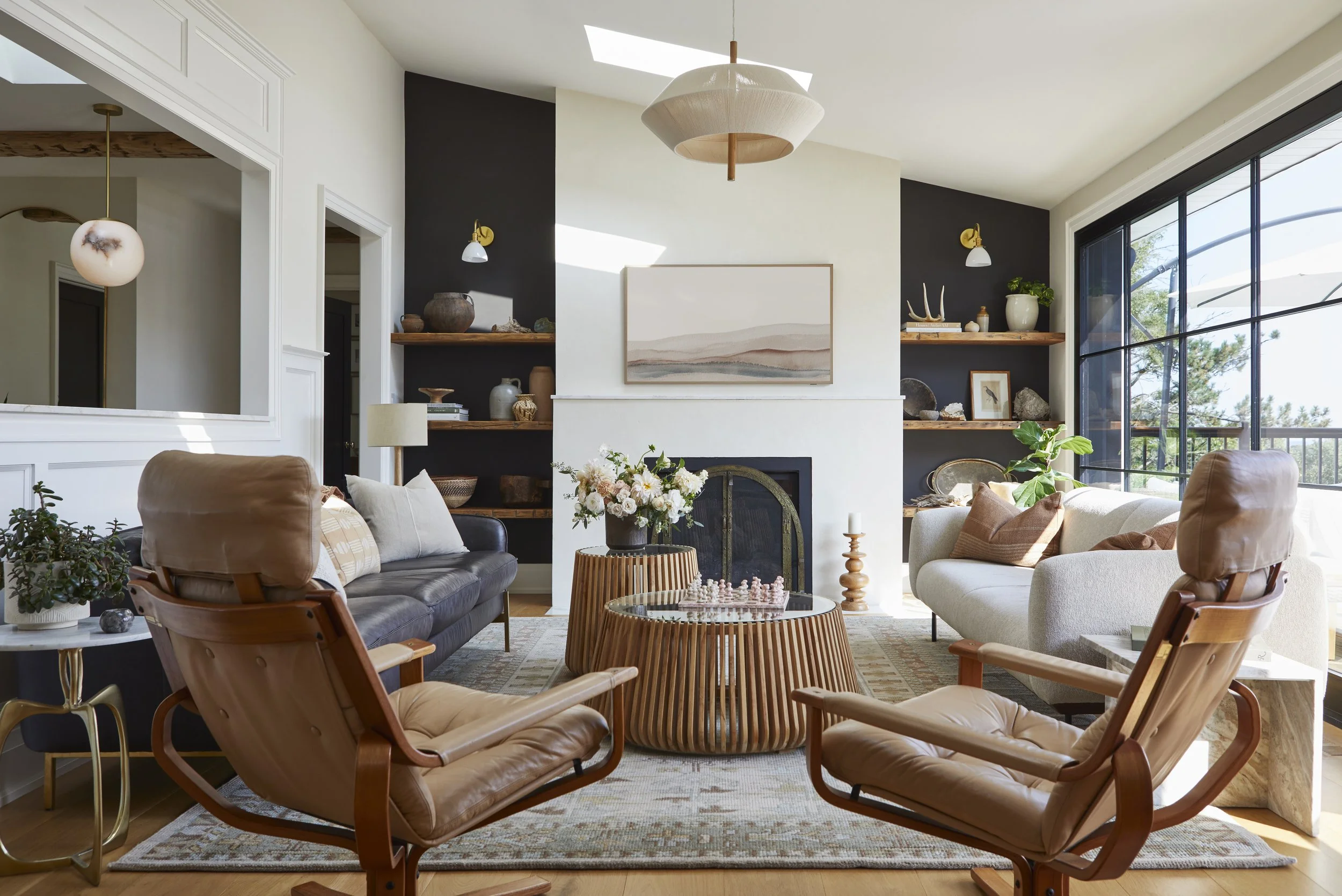 Living room with two beige armchairs, a white sofa, a black leather sofa, a fireplace, wooden shelves with decorative items, large windows, and a central coffee table with a flower arrangement