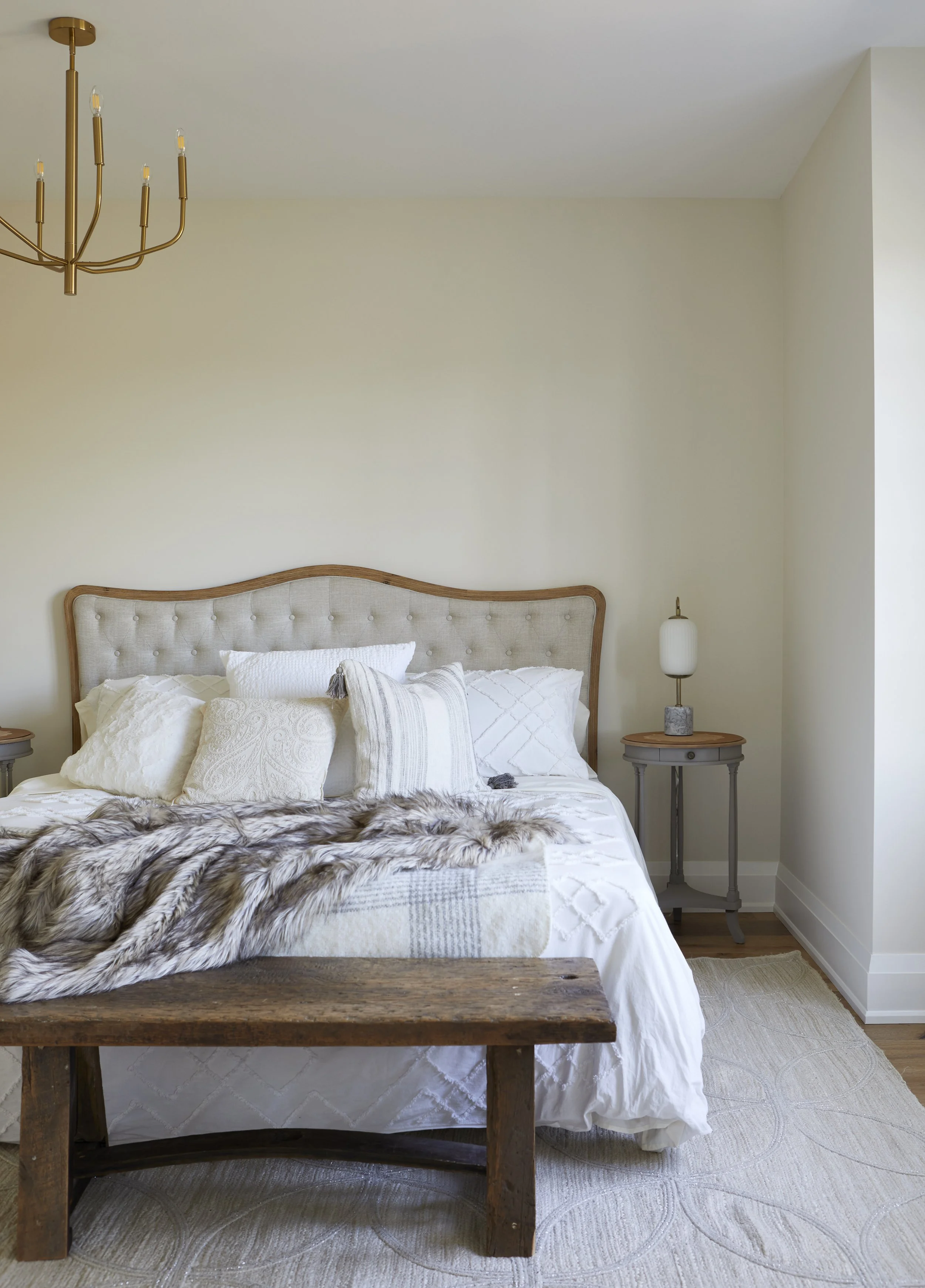 A bedroom with a beige tufted headboard, white bedding, and multiple pillows. There is a fur throw and a wooden bench at the foot of the bed. On each side of the bed, there are small gray nightstands; the right one has a lamp with a white shade. A go