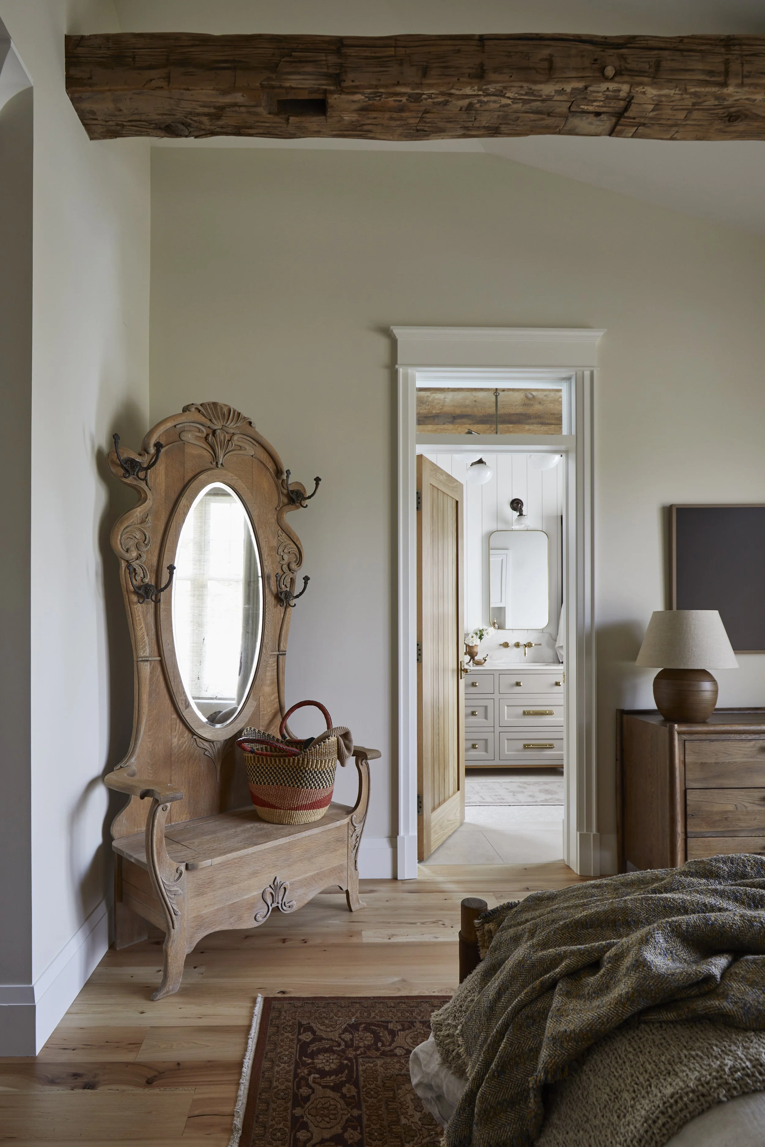 A rustic bedroom with wooden accents, featuring a vintage wooden hall tree with an oval mirror and hooks, a woven basket on it, and a bed with a textured blanket. A doorway leads to a bright bathroom with white cabinetry and a mirror.