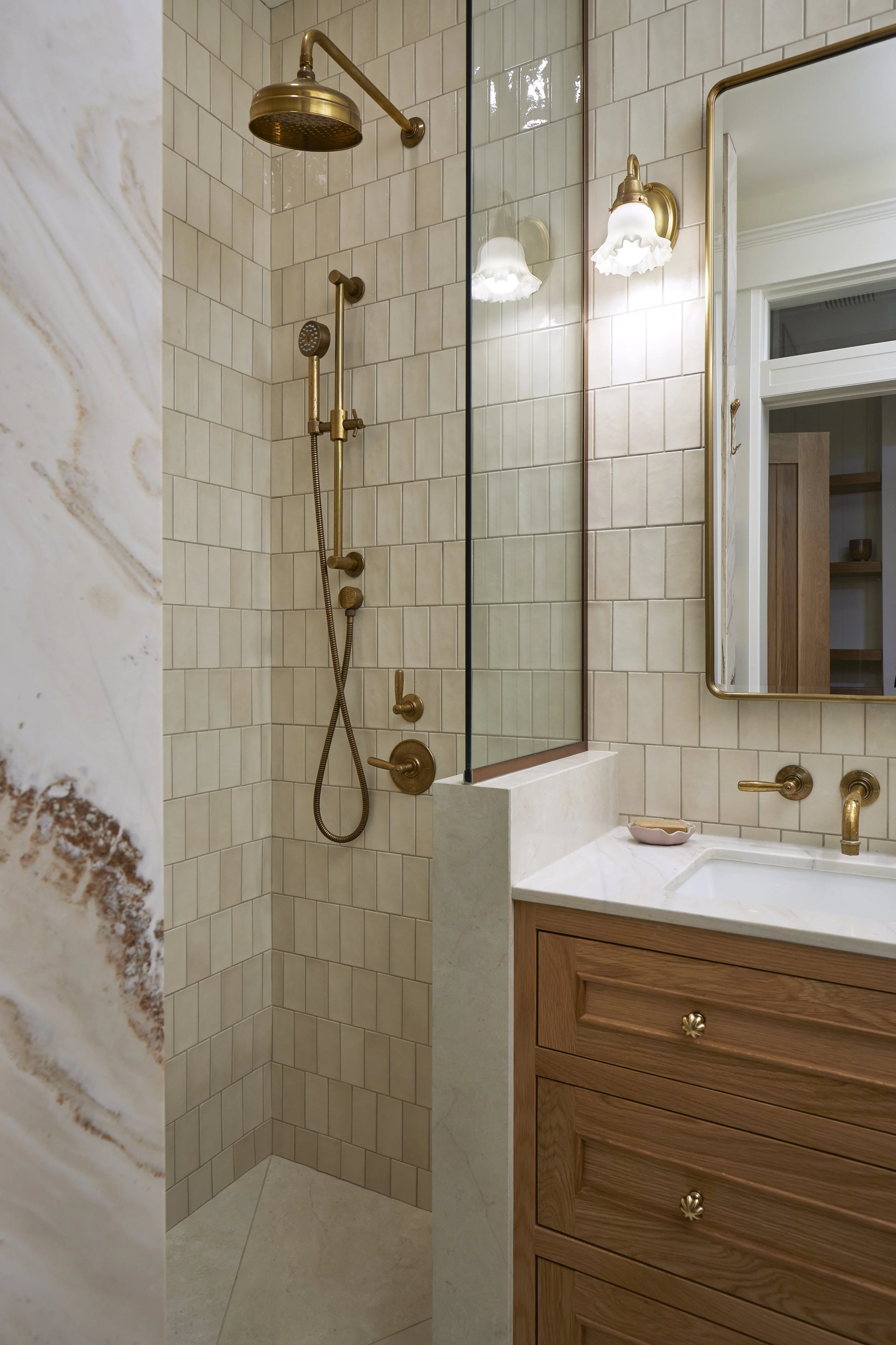 A bathroom with a walk-in shower, featuring a brass showerhead, handheld shower, and fixtures, beige tiled walls, a wooden vanity with a white marble countertop, and a mirror with brass framing.