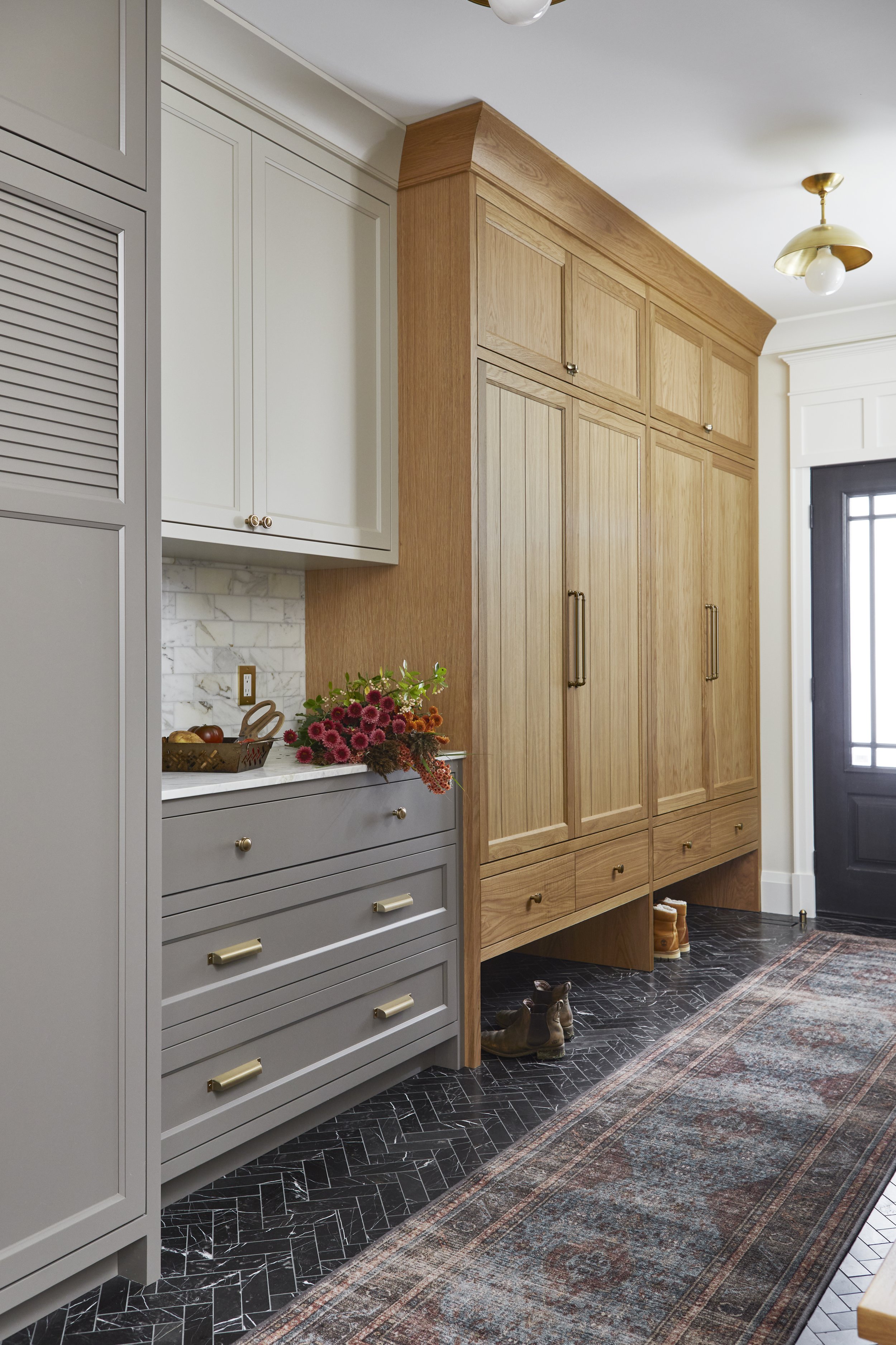 Entrance hallway with a black door, wood and gray cabinetry, a floral arrangement, and a patterned rug on black herringbone tile.