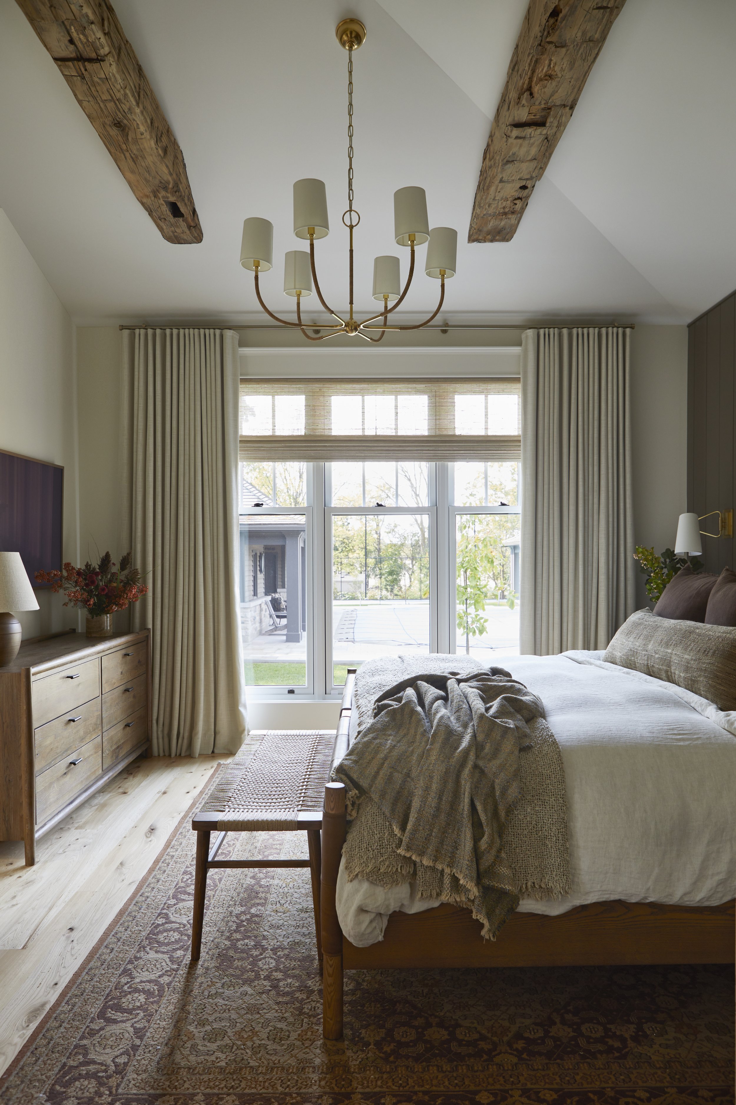A cozy bedroom with a large bed, wooden furniture, a chandelier, a window with curtains, and a view of a backyard.
