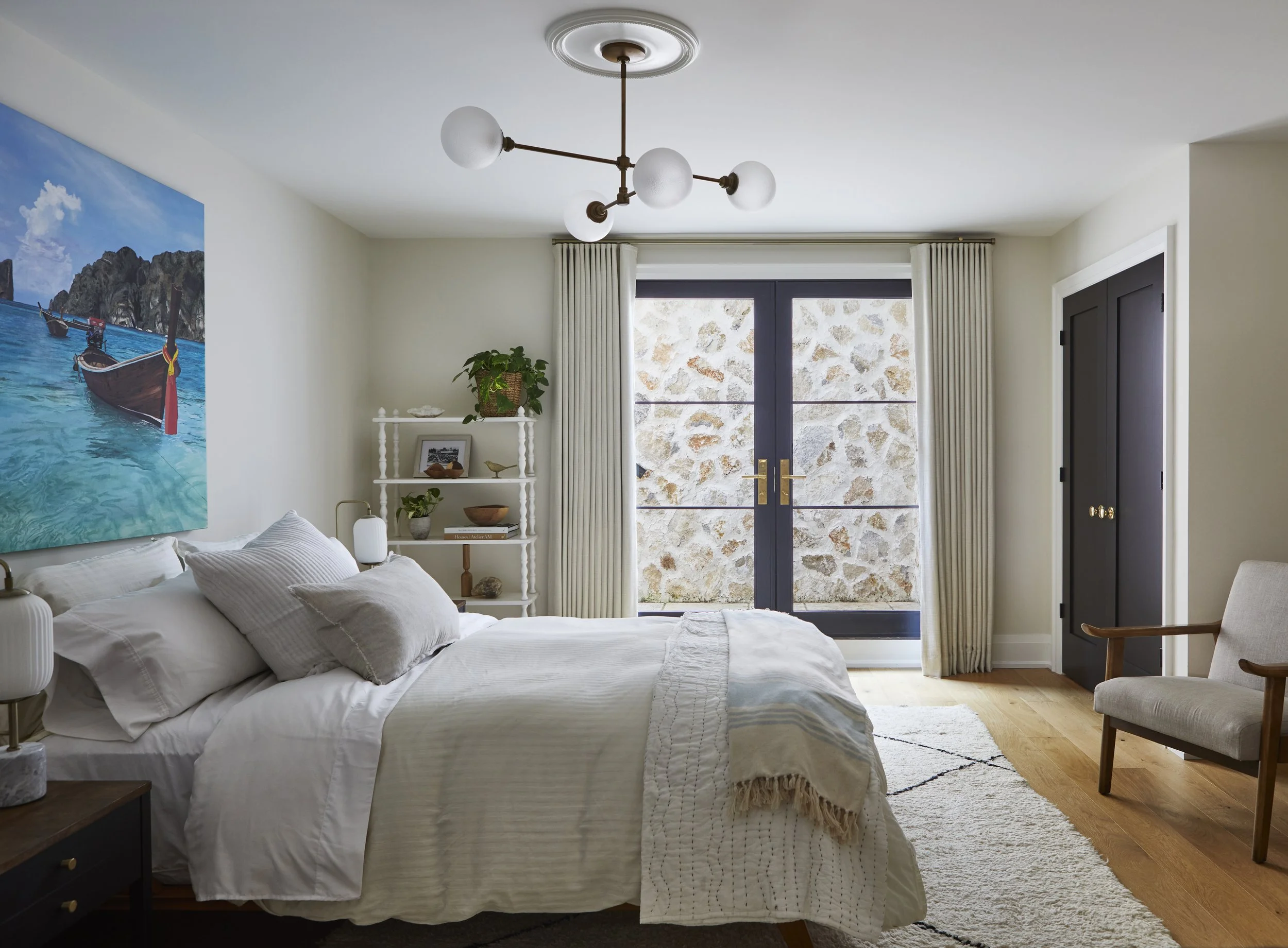 A cozy bedroom with a white bed, large window with curtains, a white bookshelf with plants and decor, a black closet, a beige armchair, and a modern ceiling light fixture.