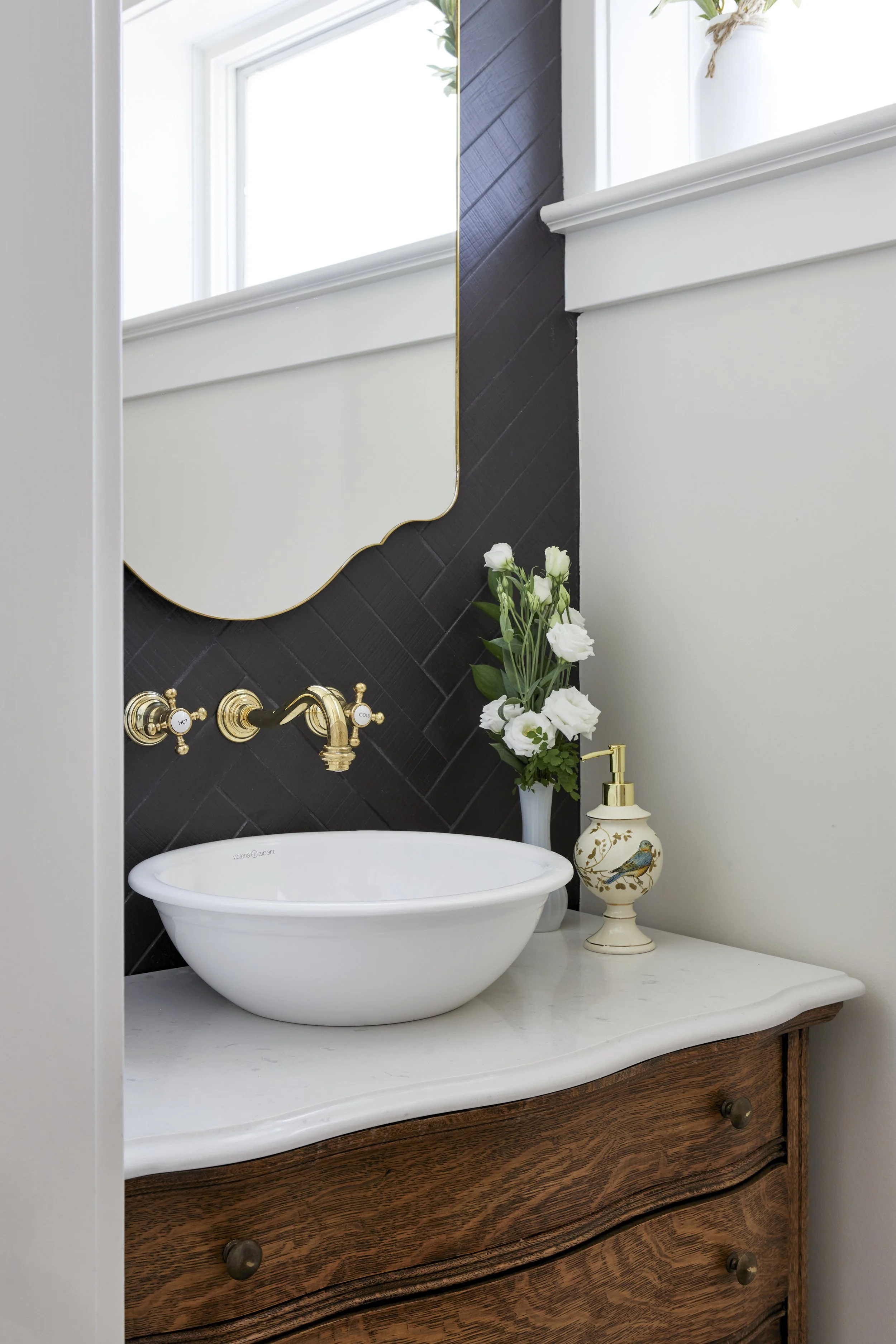 Bathroom vanity with a white vessel sink, gold fixtures, a mirror, a vase with white flowers, and a decorative soap dispenser against a white wall and black tiled accent wall.