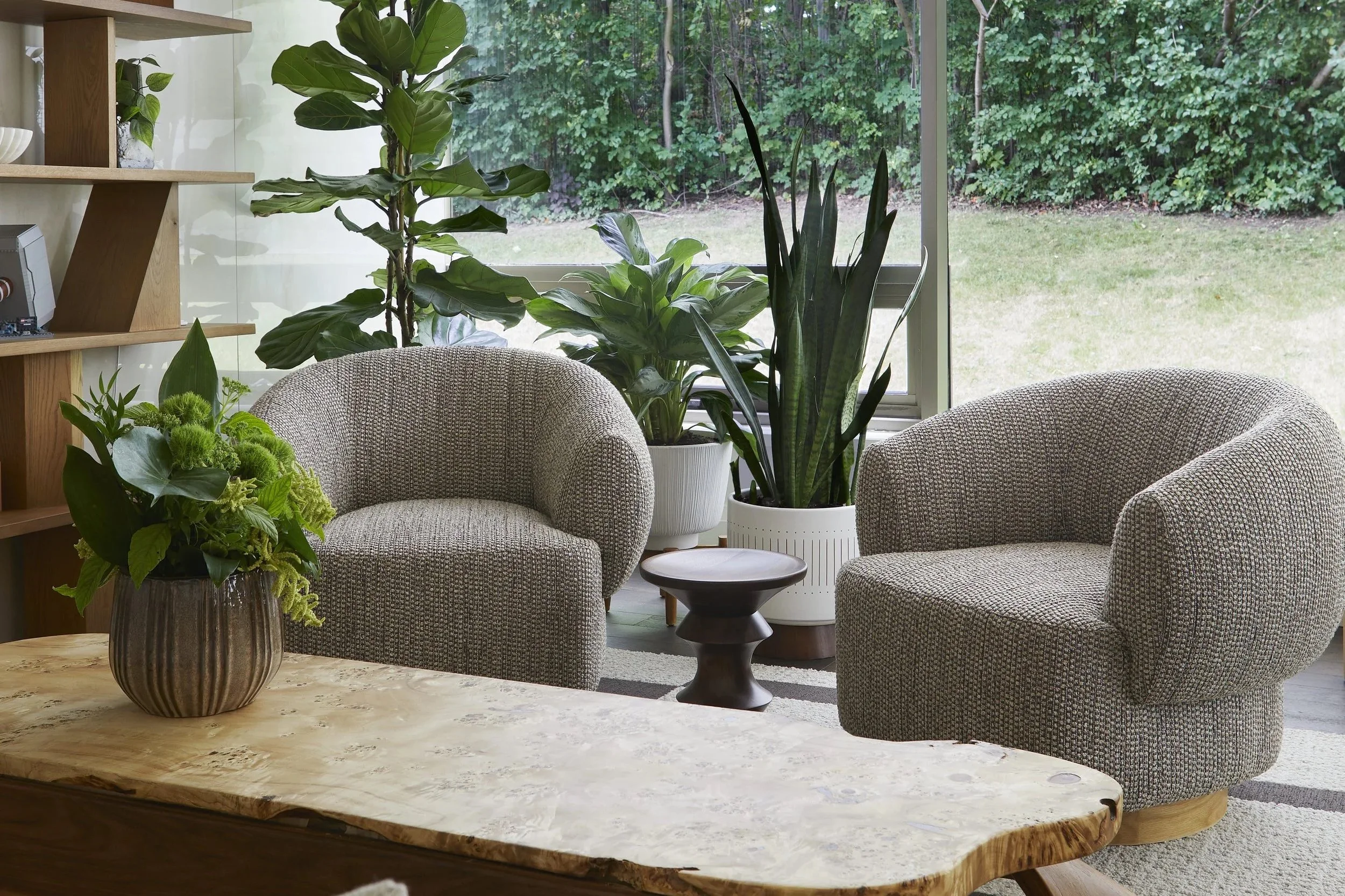 Living room with two upholstered armchairs, large potted plants, a wooden coffee table, and a large window overlooking a green backyard.