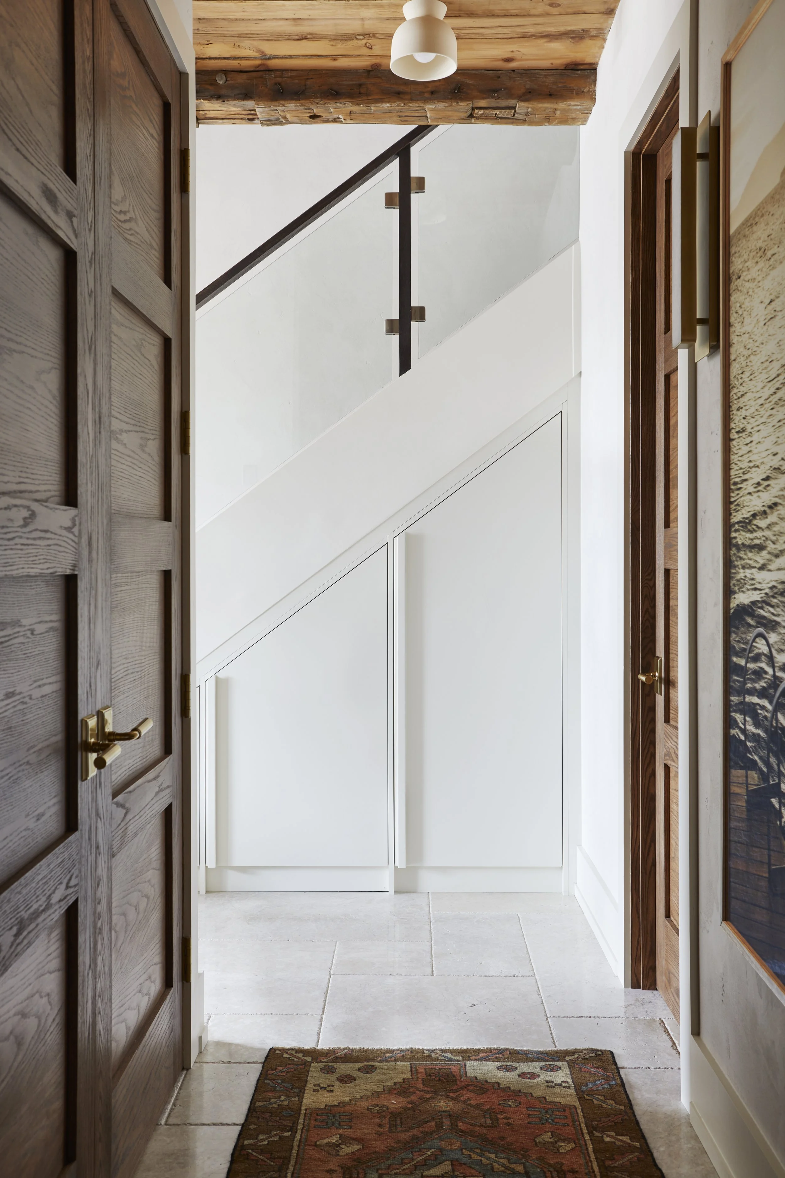 Interior looking toward staircase with glass and black railing, white paneled wall, wooden doors, and a patterned rug on light-colored tile flooring.