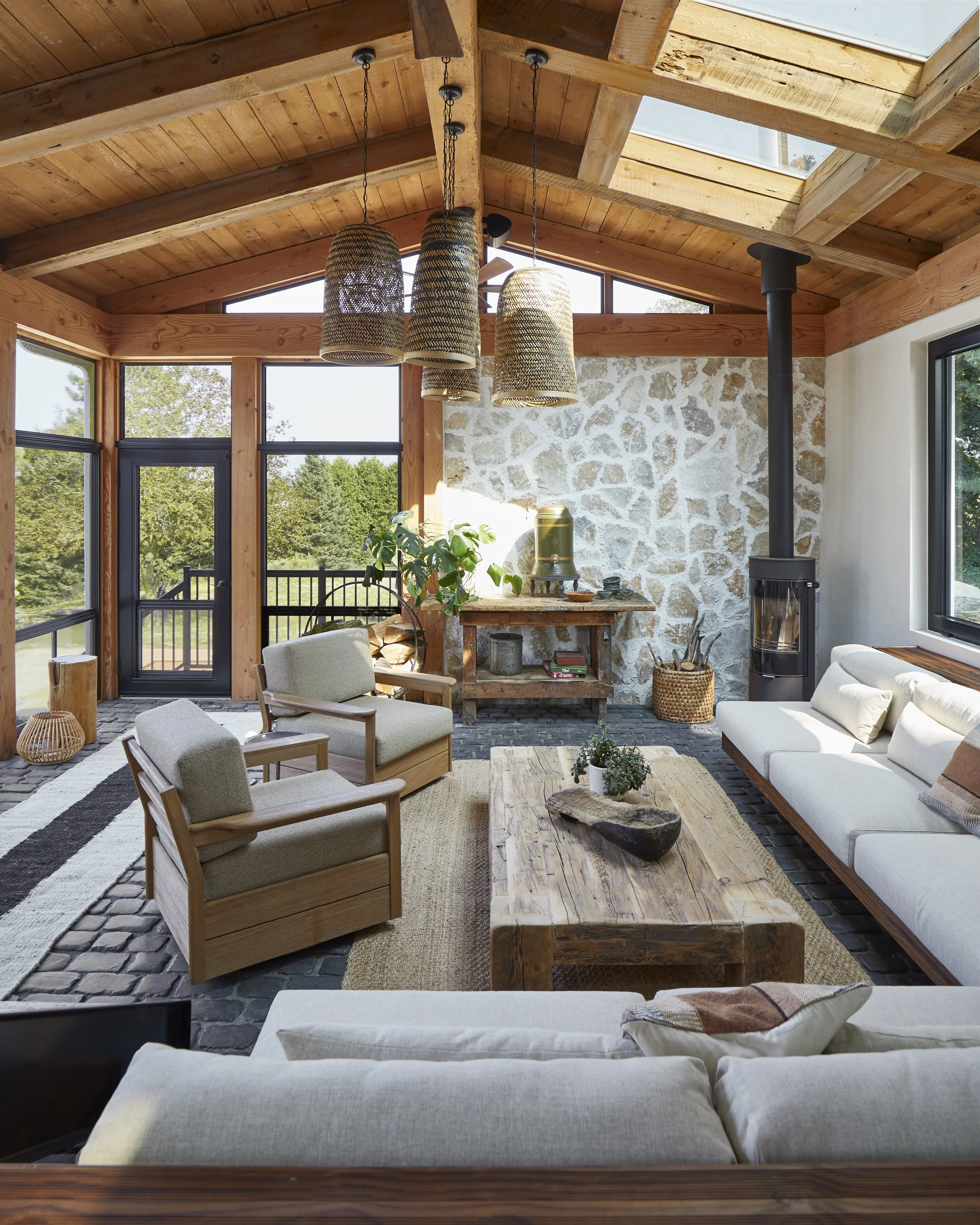 Cozy living room with large windows, wooden ceiling, and stone wall, featuring white sofas, wooden armchairs, a rustic coffee table, and a wood stove.