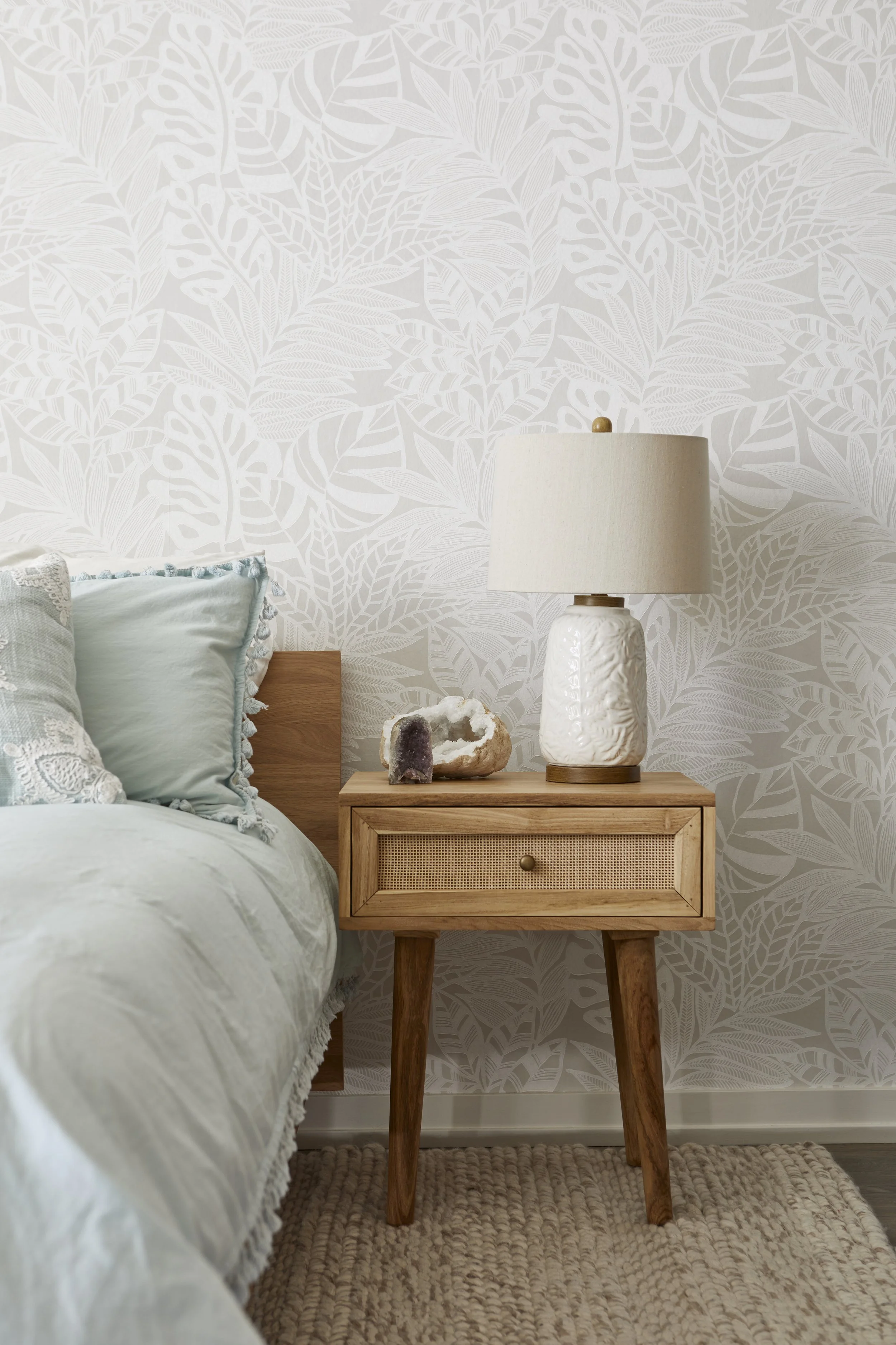 A bedroom with a wooden nightstand, a ceramic table lamp, a purple and white geode, and a white decorative mineral specimen. The wall behind has a leaf-patterned wallpaper, and there are pillows with lace trim on the bed to the left.