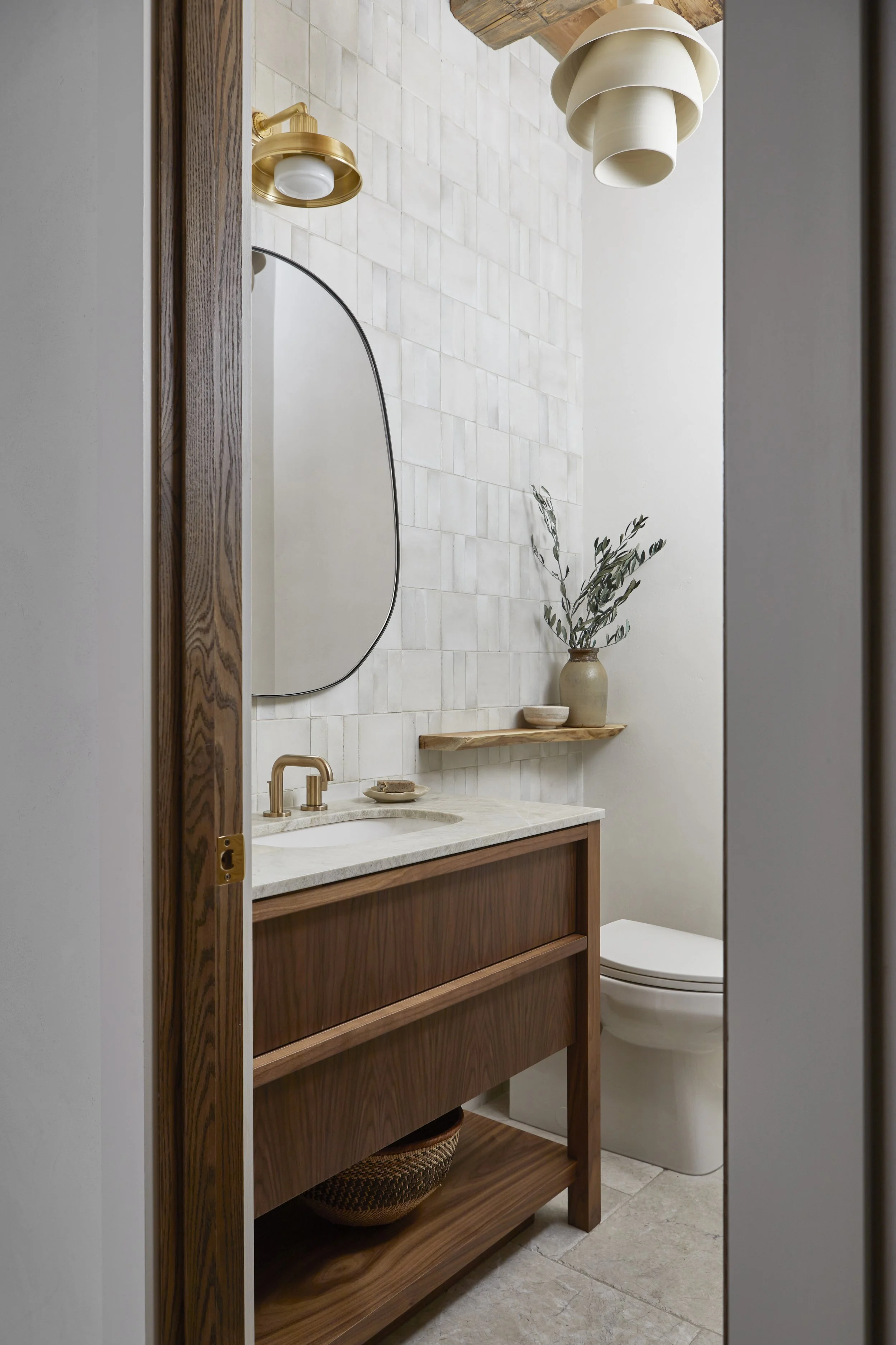 A small, modern bathroom with a wooden vanity, a white oval toilet, a mirror, a potted plant, and a unique layered ceiling fixture.
