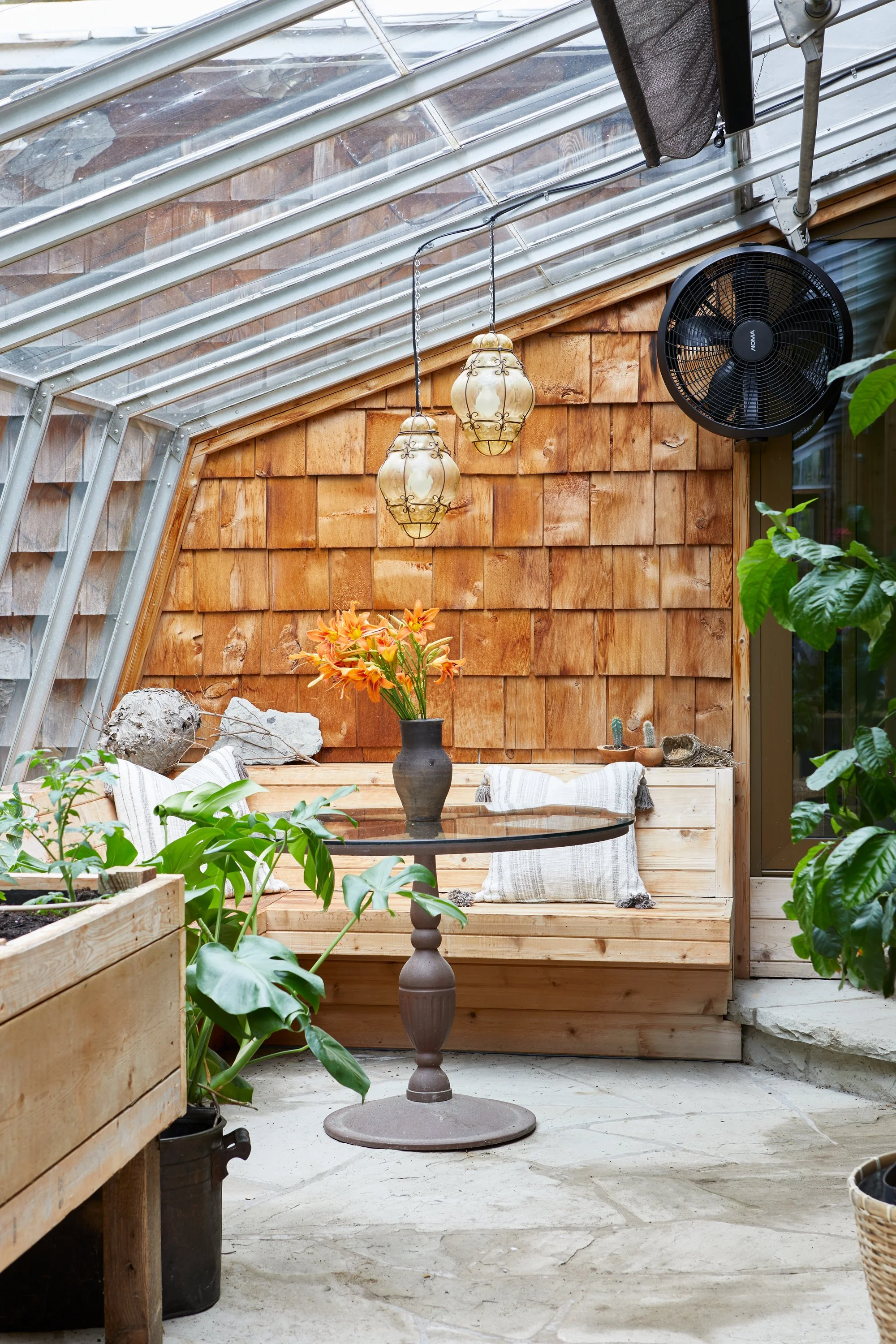 Indoor greenhouse with wooden walls, glass ceiling, potted plants, a round table with a vase of orange lilies, hanging pendant lights, and a black fan.