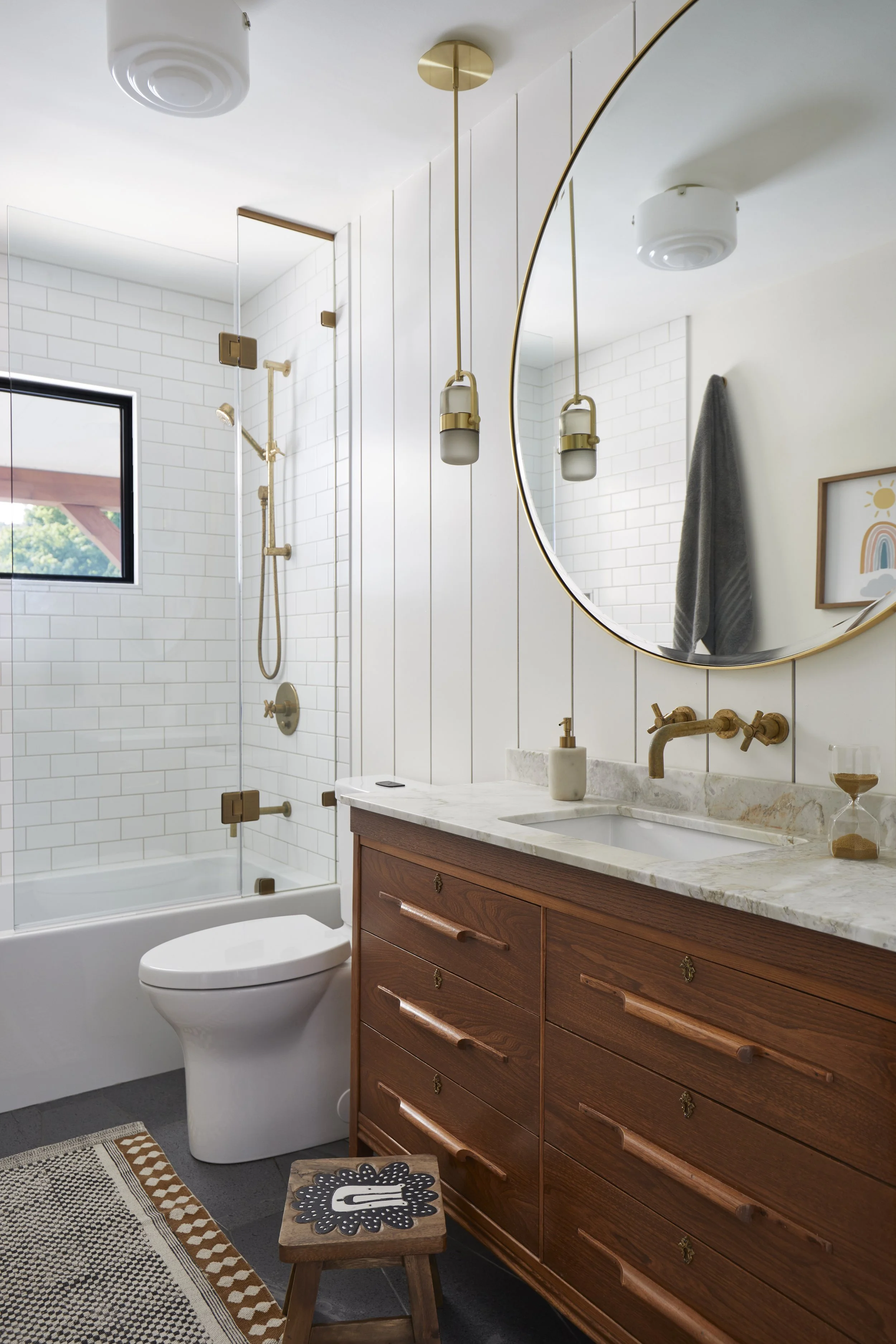 Modern bathroom with white subway tile walls, a shower with gold fixtures, a round mirror, and a wooden vanity with a white marble countertop.