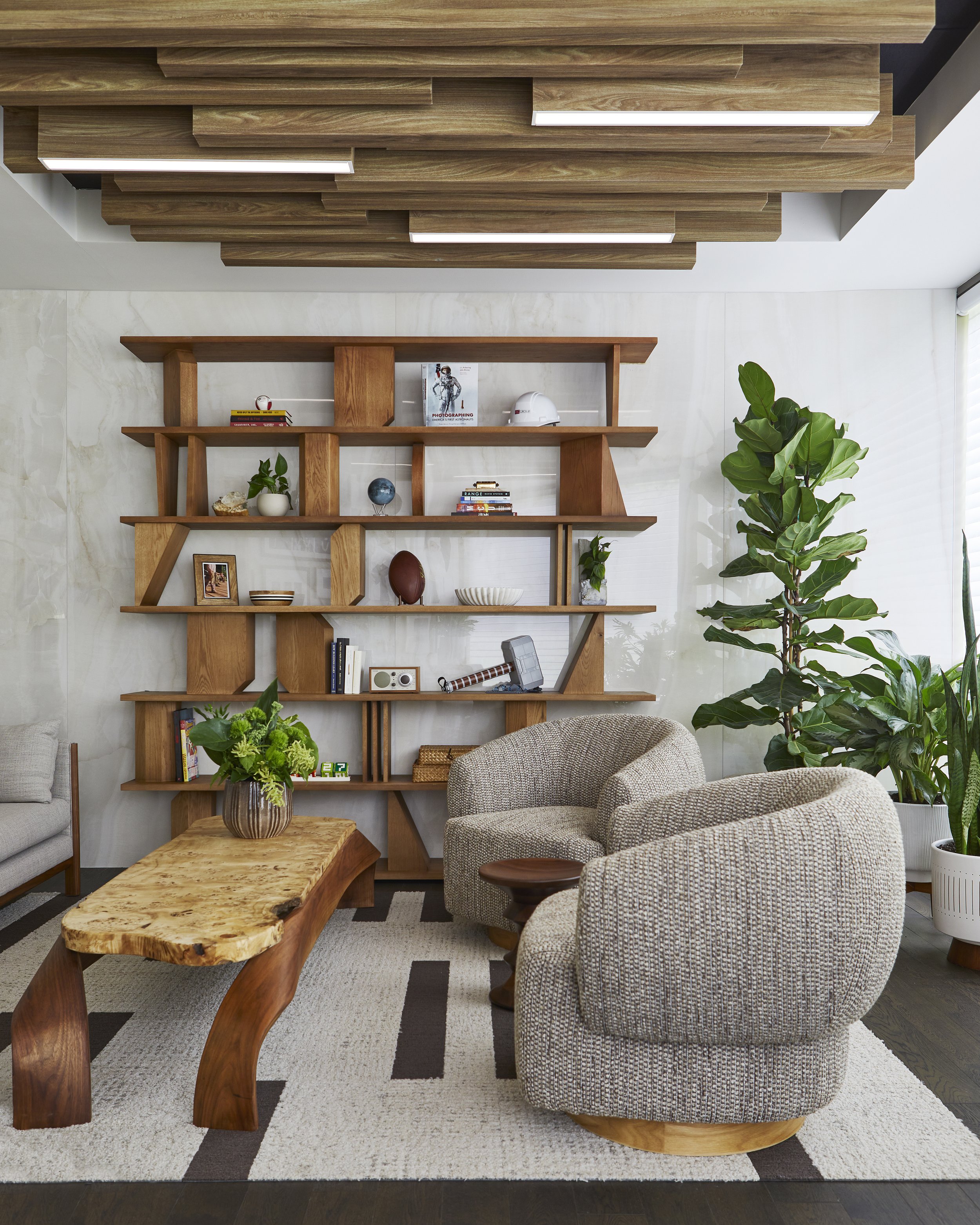 Living room with a beige patterned armchair, a unique wooden coffee table with a natural finish, a large wooden bookshelf with various decor items, and several green potted plants near a window. The room has a light-colored wall, a textured white and