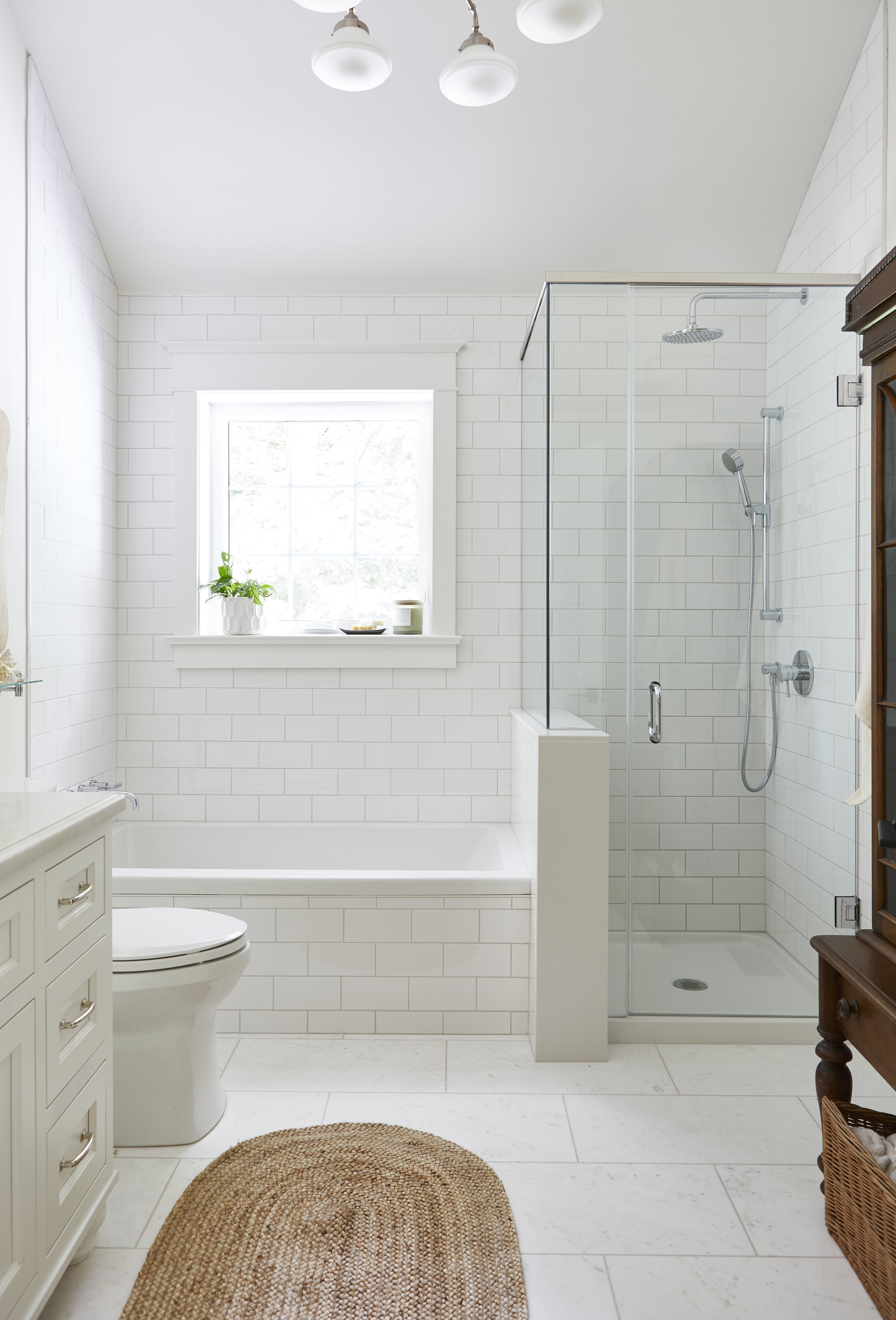 A clean, white bathroom with a window, a bath, a shower with glass door, and a brown wooden cabinet.