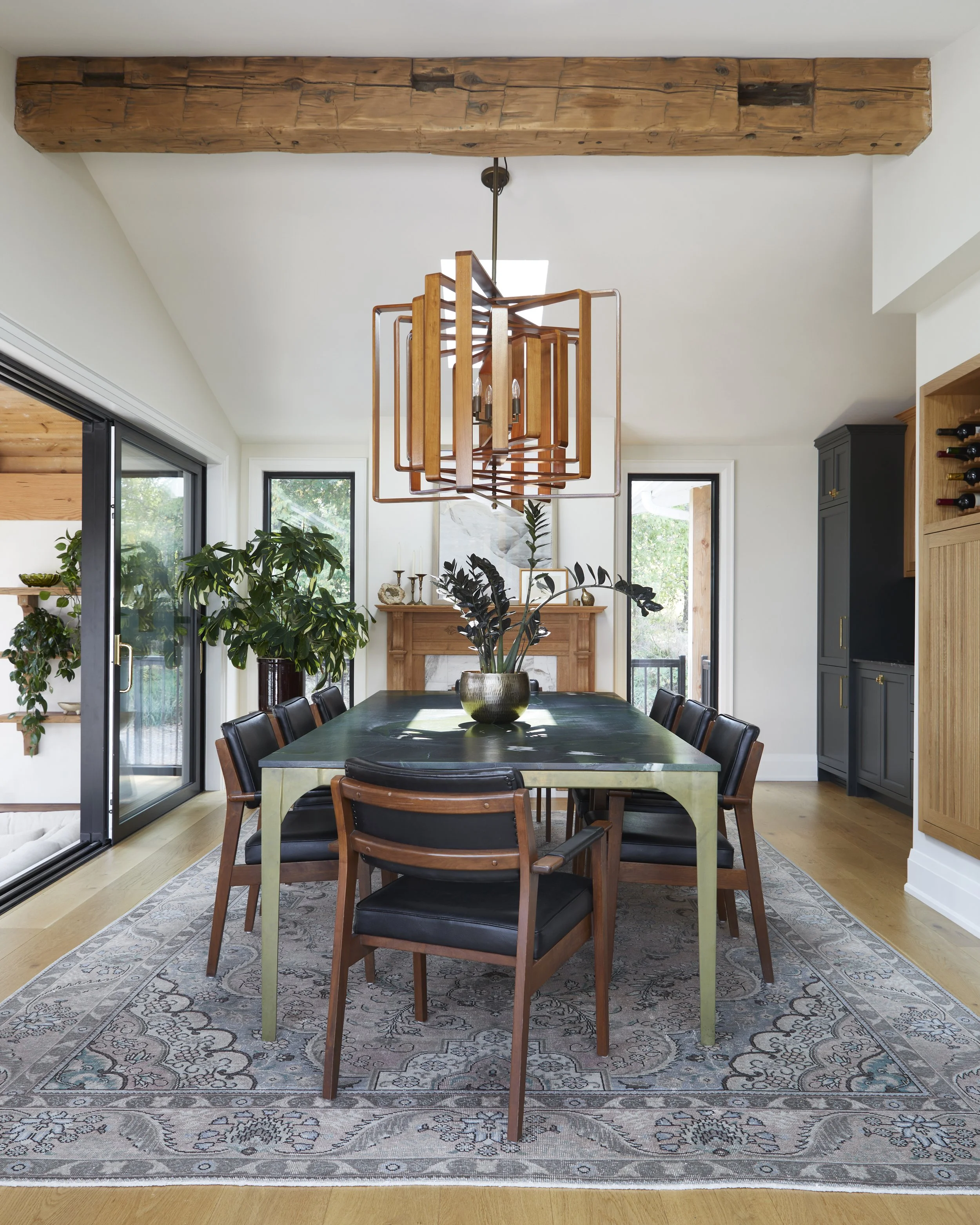 A dining room with a large dark green table, black and wooden chairs, a decorative plant centerpiece, a wooden beam on the ceiling, a unique wooden chandelier, a fireplace, and large windows with outdoor views.