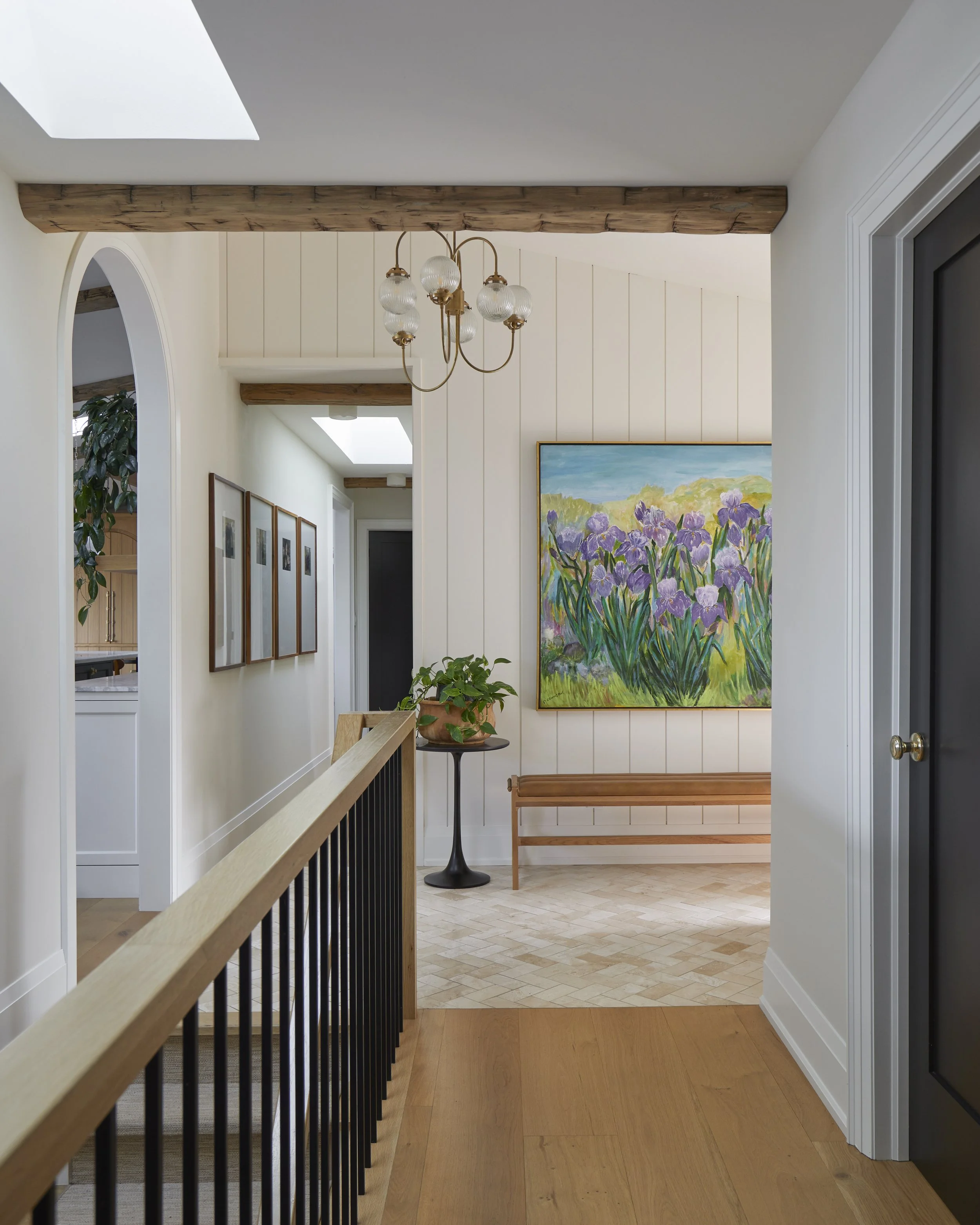 Bright hallway with a chandelier, a large colorful floral painting, a bench, a potted plant on a stand, wood beams, and wood flooring.