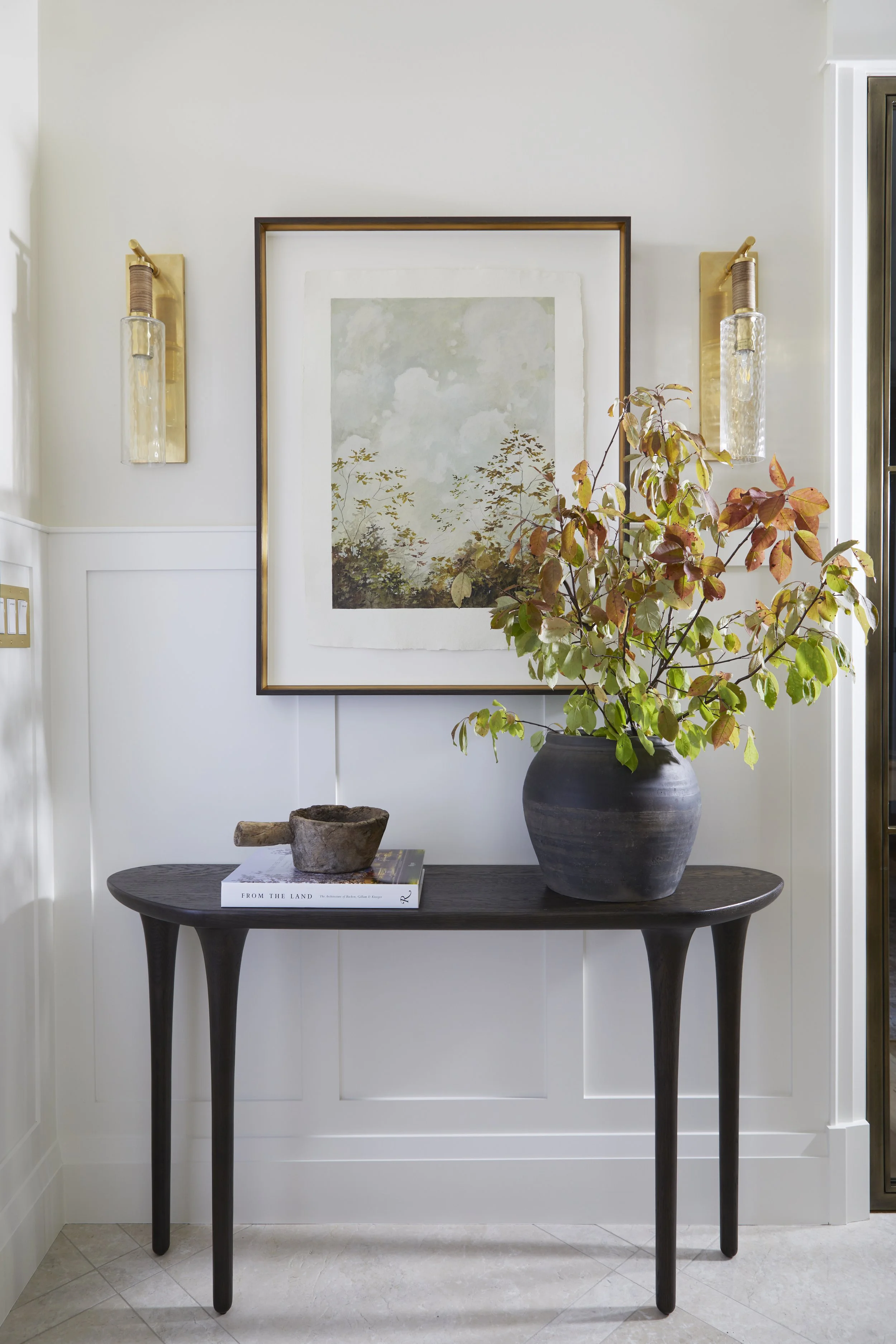 A minimalist entryway with a dark wooden console table, a large black pottery vase with autumn leaves, a book and a small wooden bowl, framed artwork, and wall sconces on a white wall.