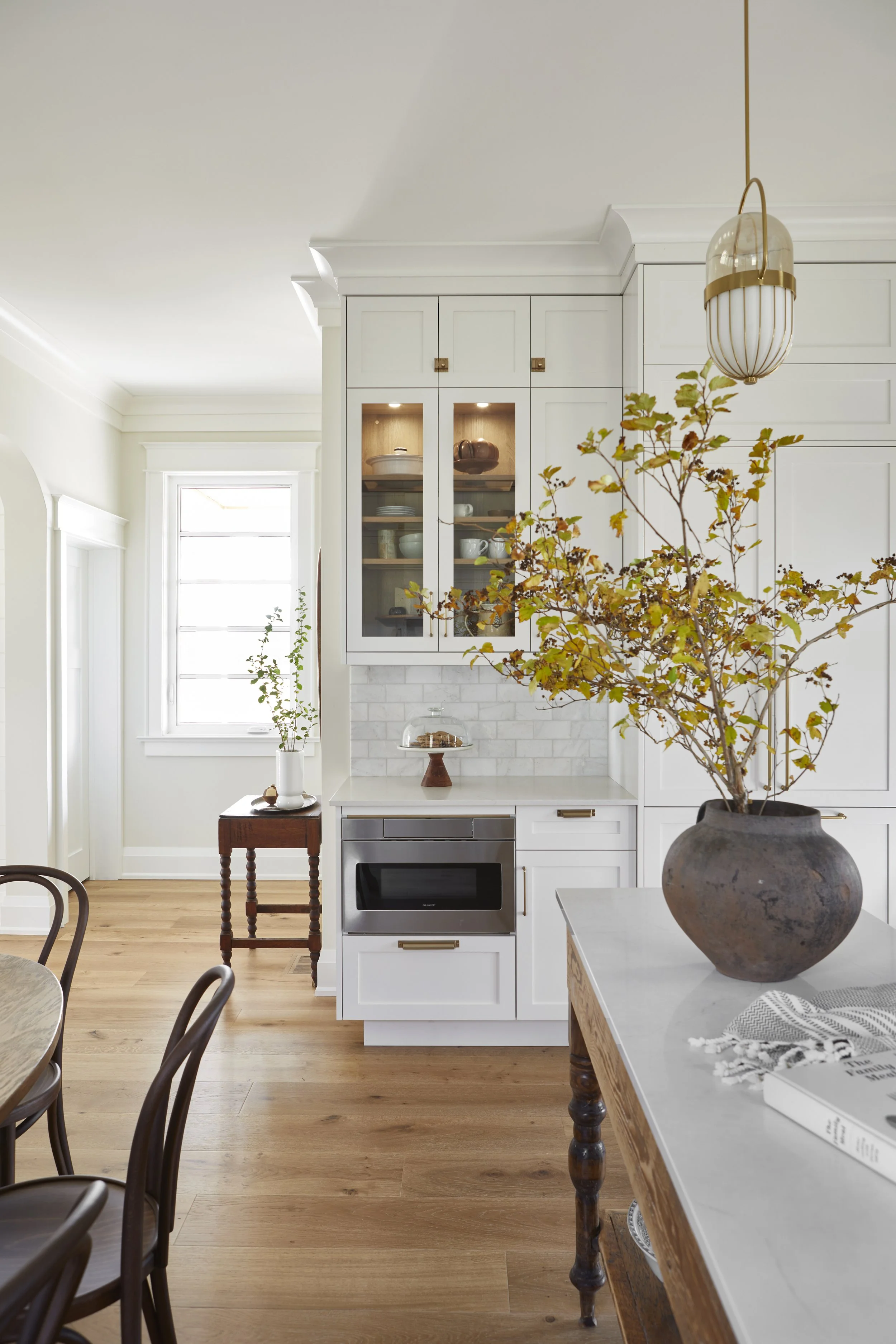 A bright, modern kitchen with white cabinetry, wooden floors, and a large table with a potted plant in a rustic vase.