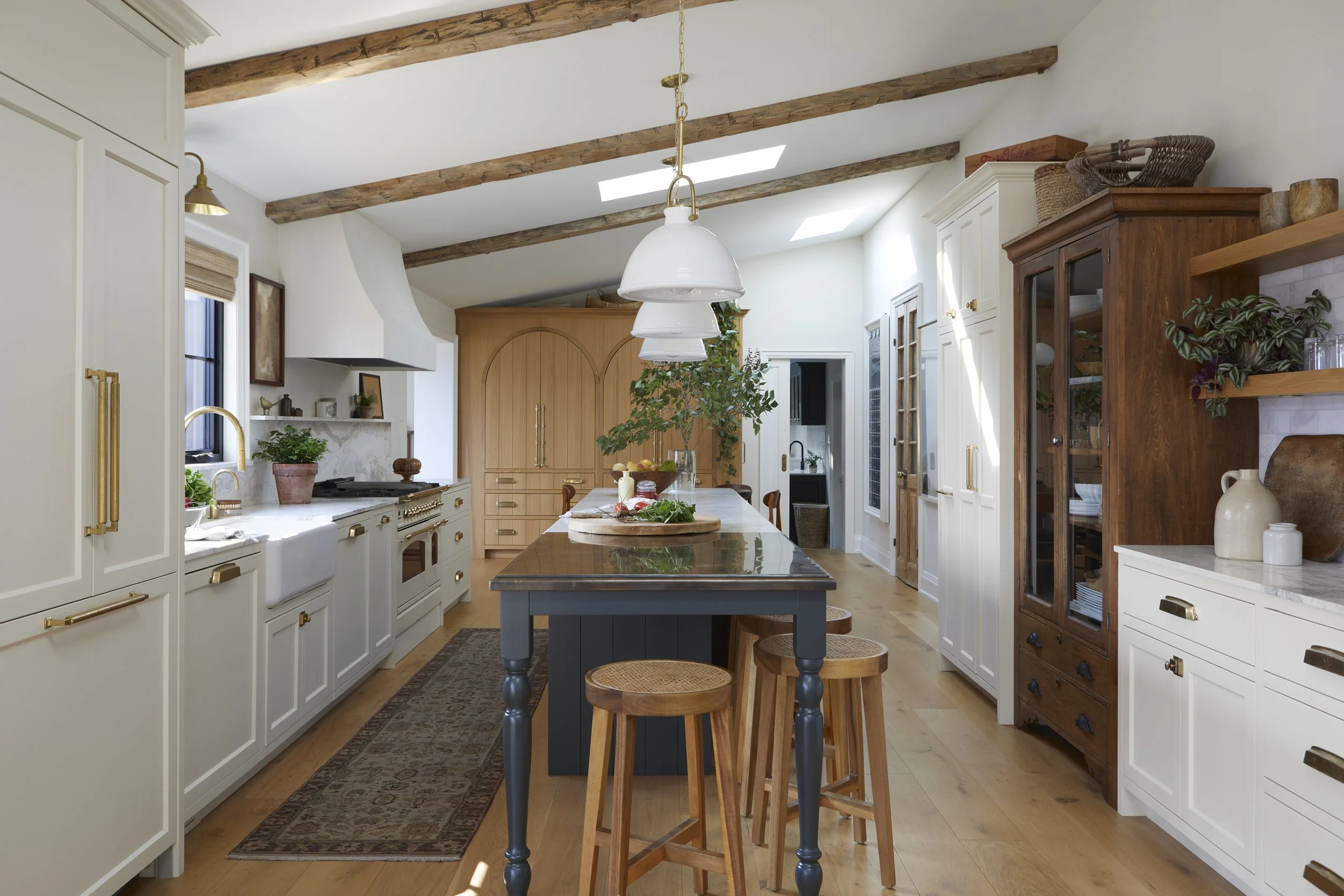 Modern kitchen with white and wood cabinetry, a central island with a dark countertop, wooden stools, and exposed wooden ceiling beams. Decor includes plants, baskets, and ceramic jars.