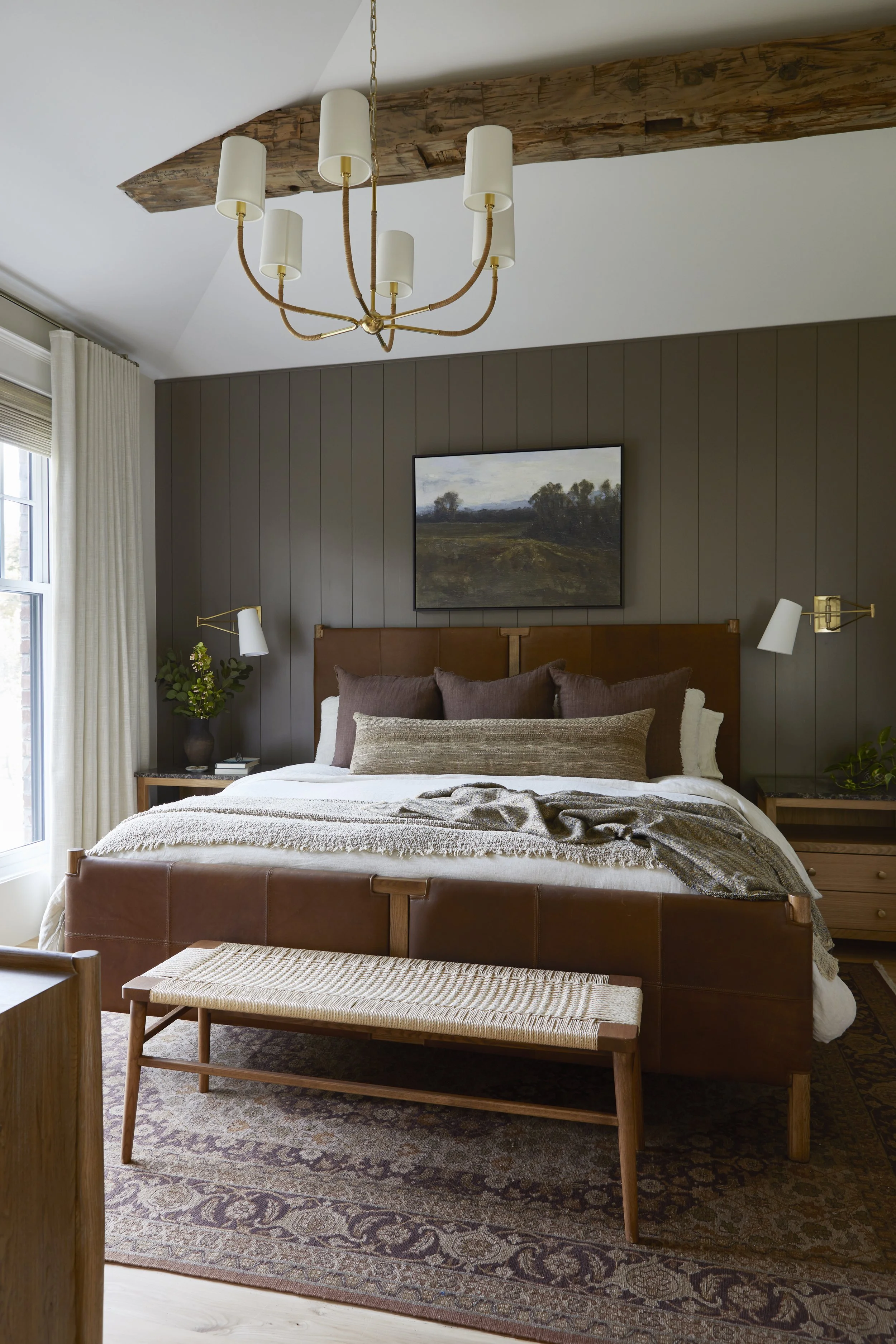 A bedroom with a brown leather bed, multiple pillows, and a textured blanket. There are two nightstands with plants and books. A picture of a landscape hangs on the dark gray paneled wall. The ceiling has a wooden beam and a modern chandelier with si