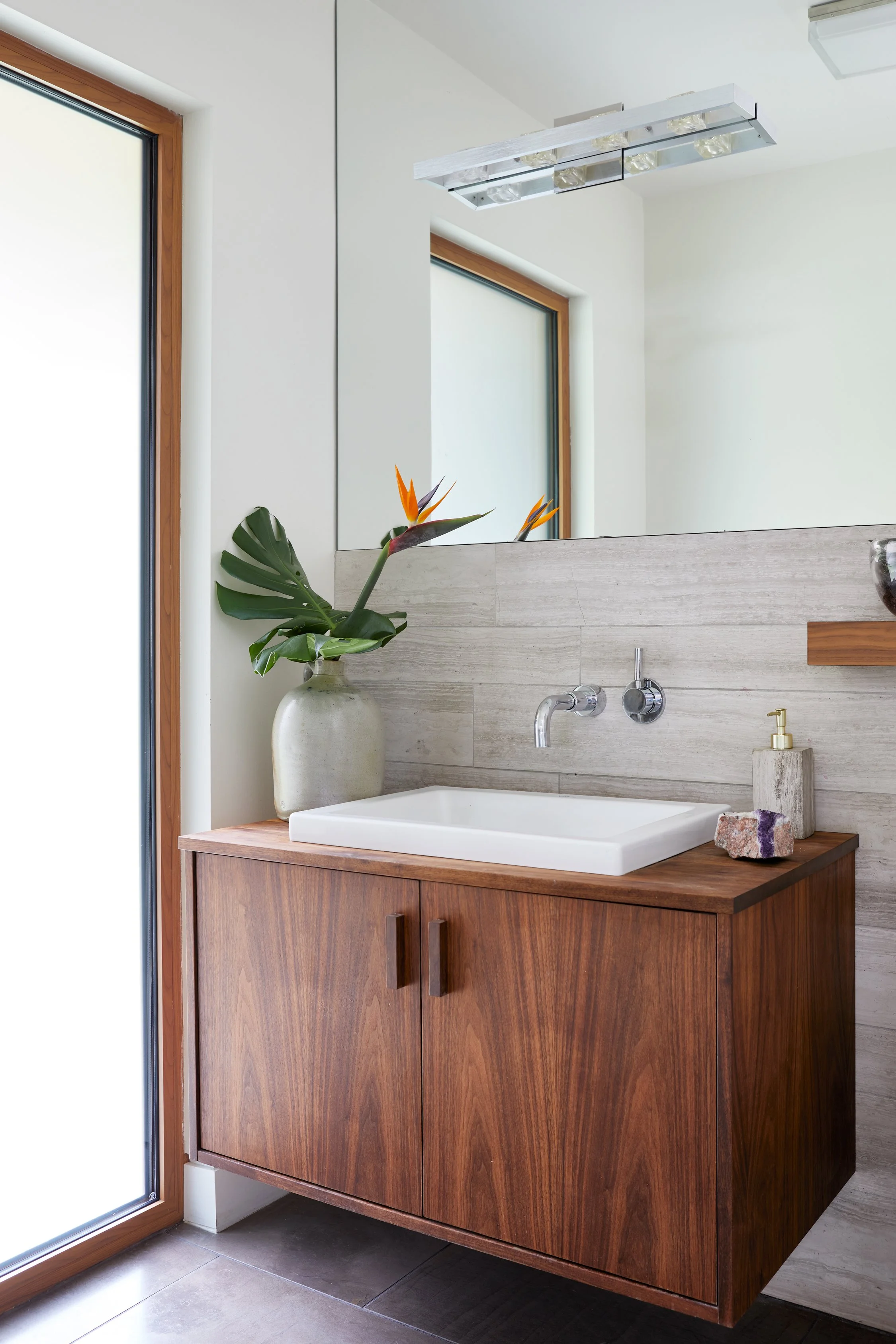 Bathroom vanity with a wooden cabinet, a white vessel sink, a wall-mounted faucet, a large mirror, a potted plant with broad green leaves and orange flowers, a soap dispenser, and a decorative stone, with a window and a modern ceiling light fixture i