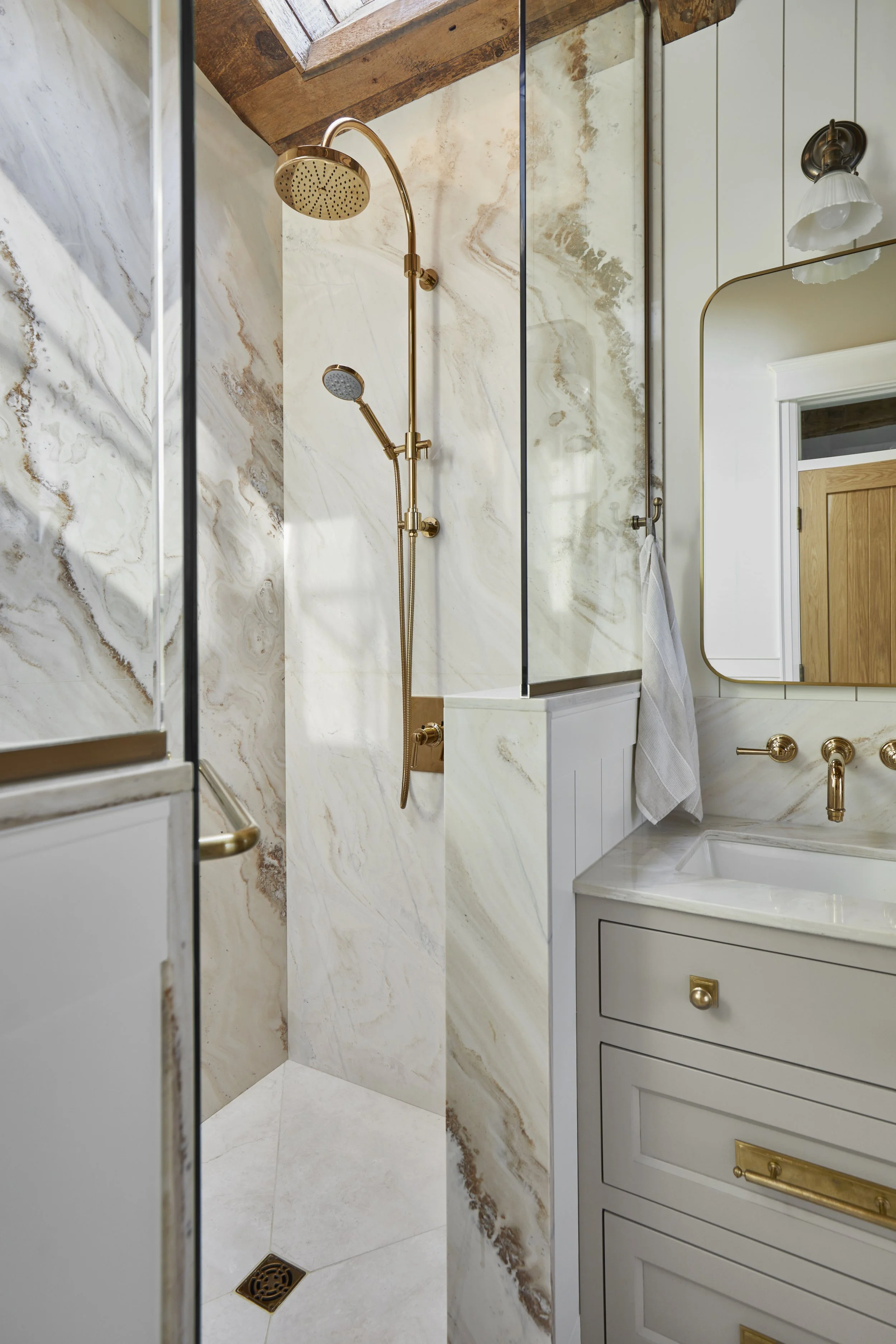 Modern bathroom with a walk-in shower featuring beige and brown marble walls and gold fixtures, next to a white vanity with gold hardware and a mirror, and a towel hanging on a gold towel bar.